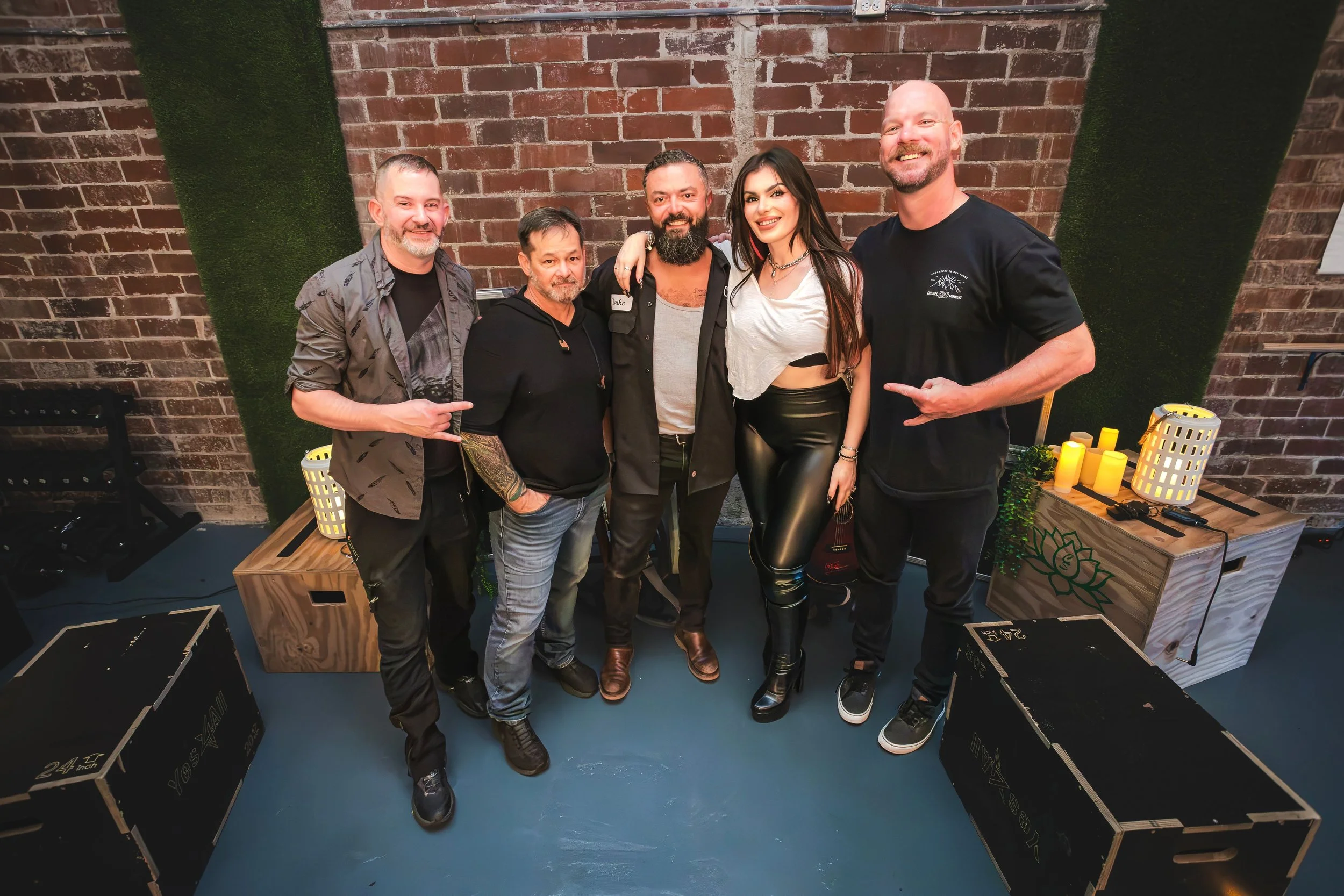 Group of six diverse people standing together indoors against a brick wall, smiling and posing for a photo, with some pointing at others, near a table with candles and boxes.