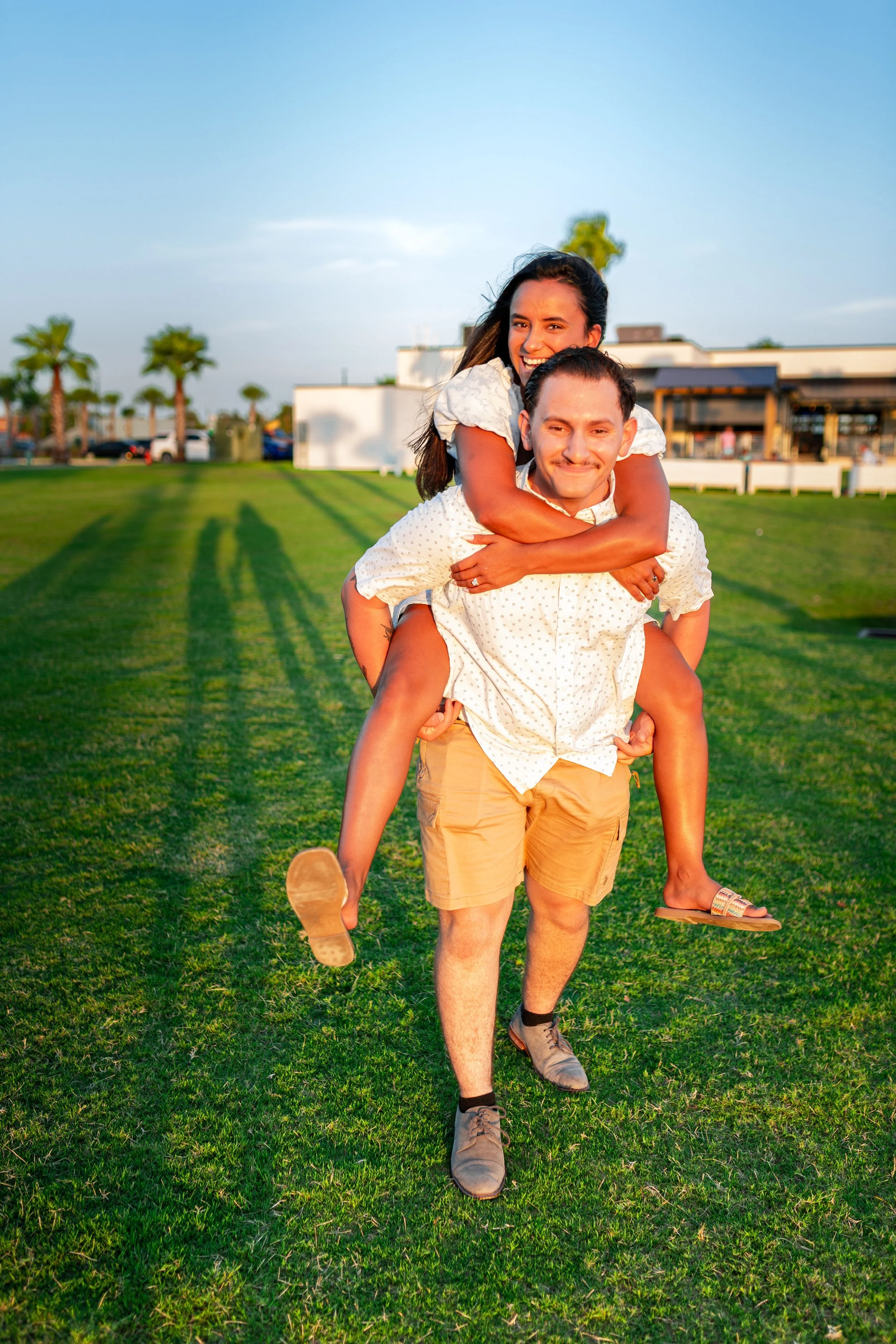 A man is carrying a woman on his back outdoors on a grassy field during sunset. The woman is smiling and holding onto the man's shoulders, while the man is smiling with closed lips. The background features palm trees, parked cars, and buildings. Both