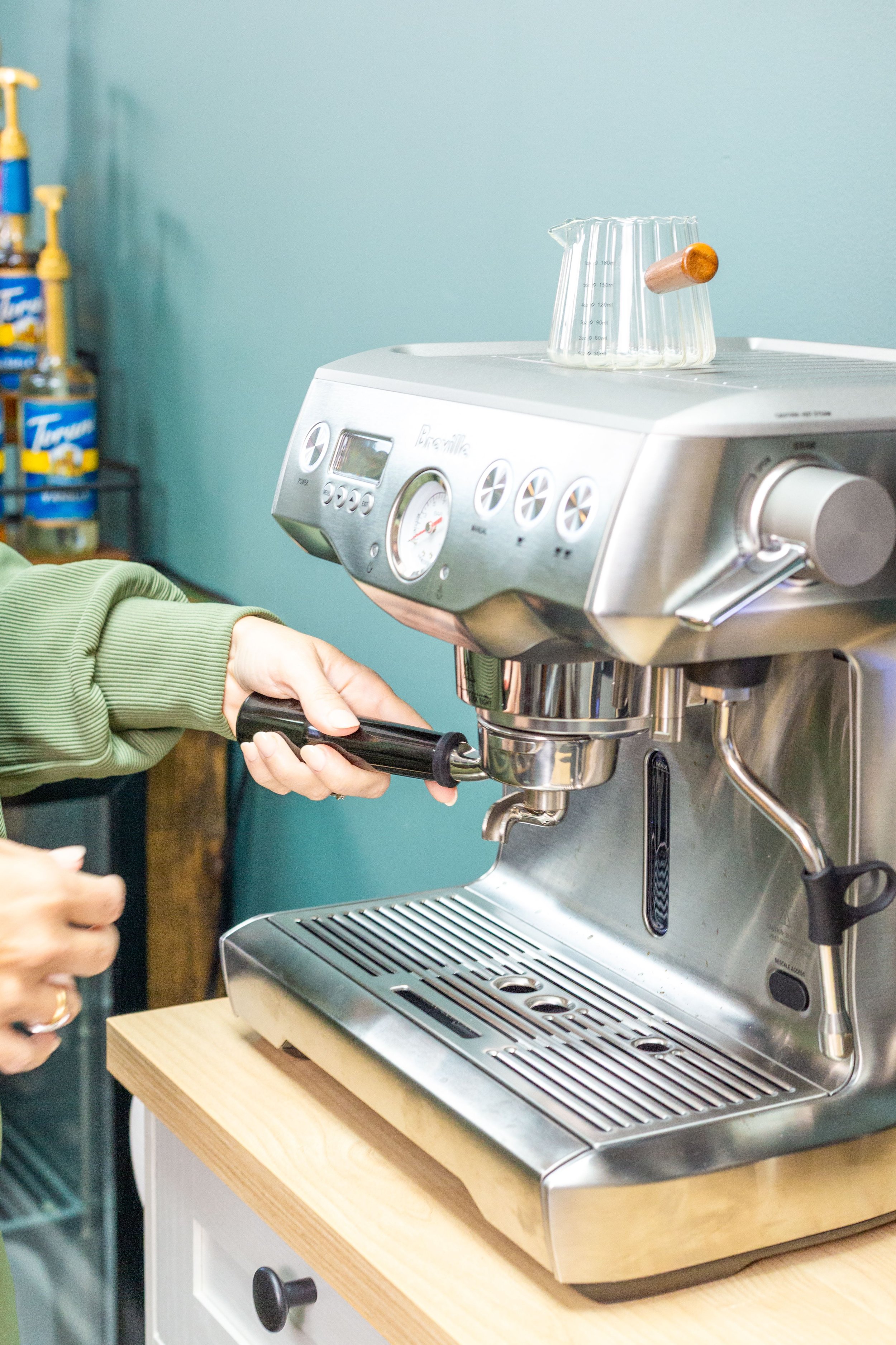 A person operating an espresso machine to prepare coffee, with a clear measuring cup on top of the machine, on a wooden countertop in a kitchen or cafe setting.