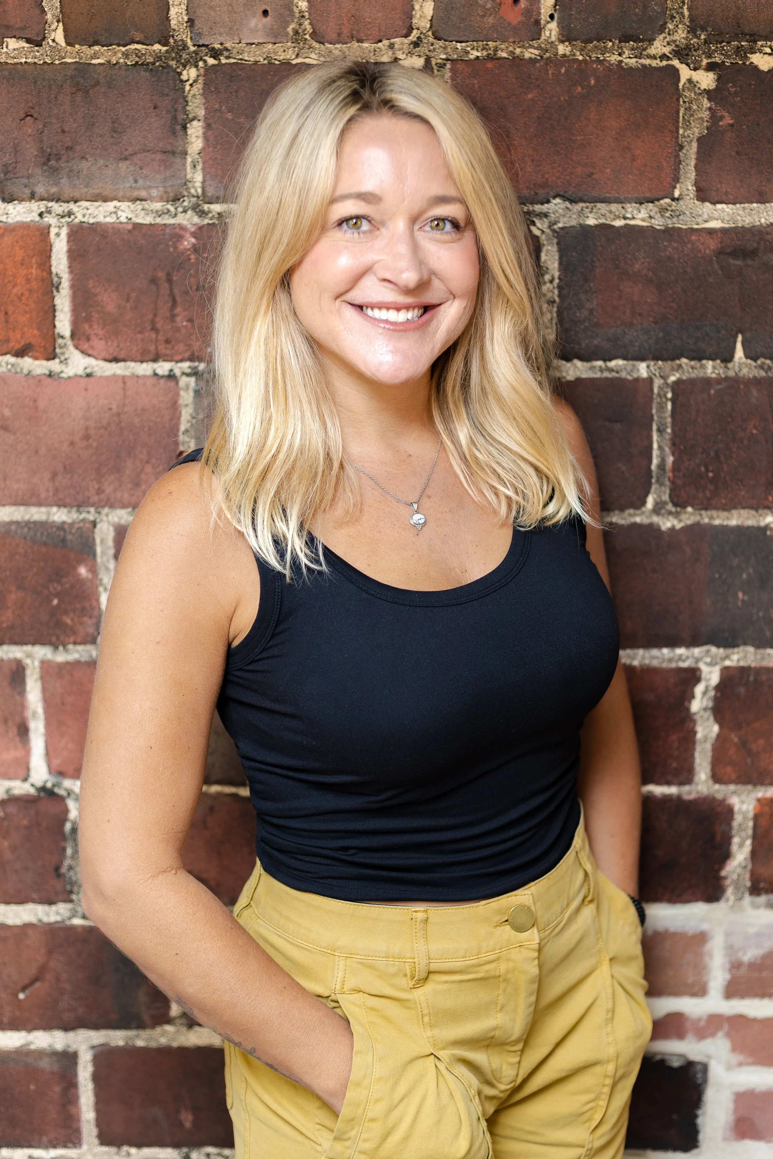 A woman with blonde hair wearing a black sleeveless top and yellow pants, standing against a brick wall, smiling at the camera.