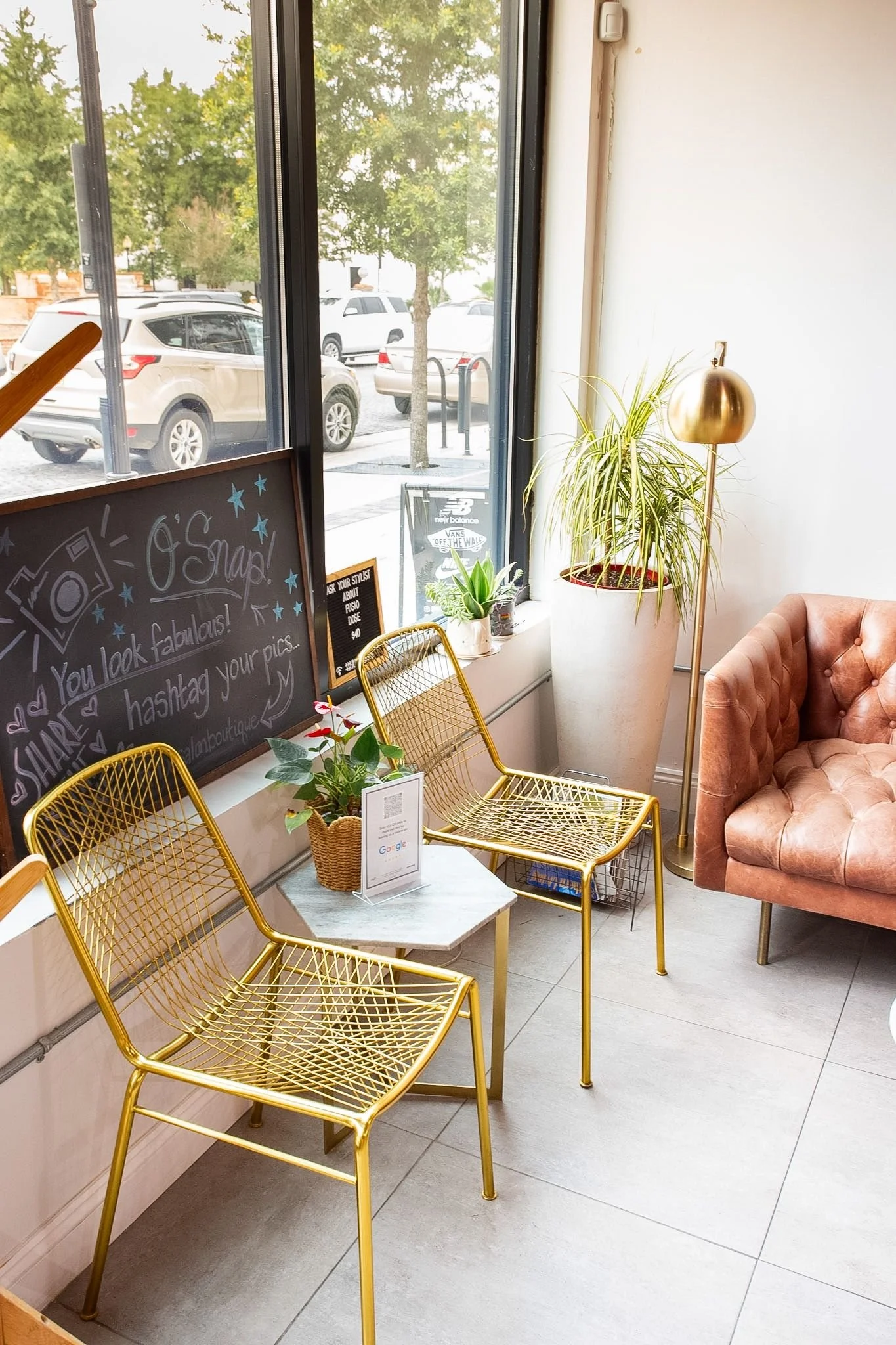Interior of a cozy cafe with gold chairs, a small marble table, a pink velvet armchair, and large windows showing parked cars and trees outside.