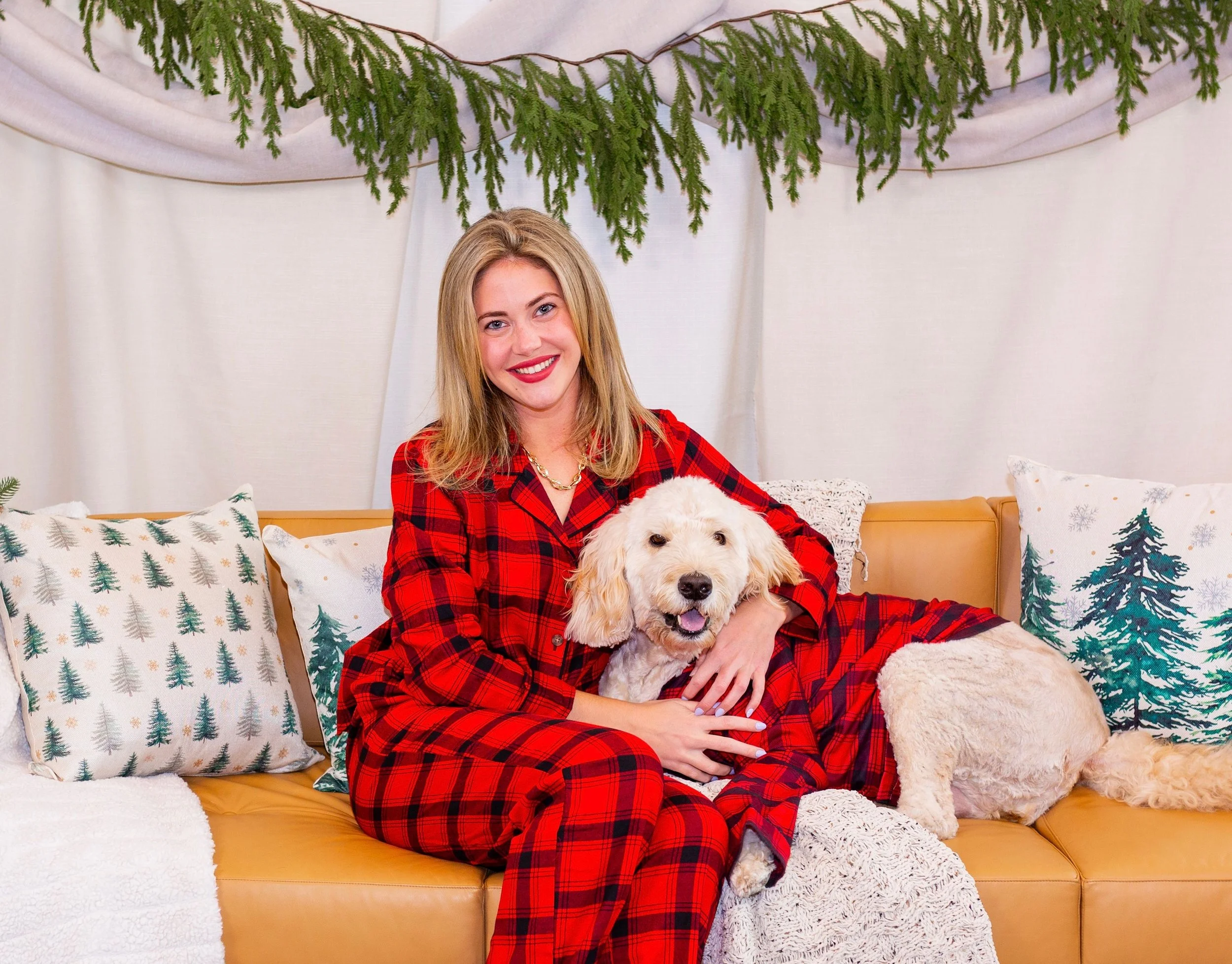 A woman with blonde hair and red lipstick smiling while sitting on a yellow sofa, holding a golden retriever puppy, in a festive decorated room with Christmas pillows, white curtains, green pine tree branches hanging from the ceiling, and a white bla
