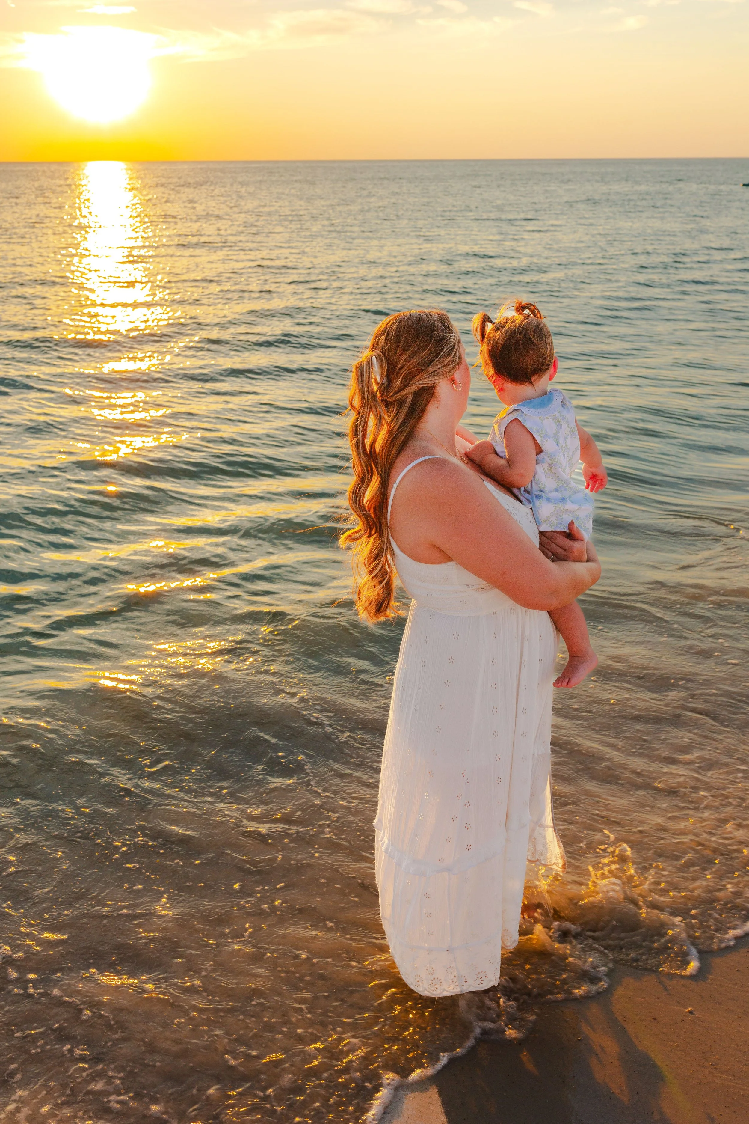 A woman in a white dress holding a young girl in a floral dress stands in shallow ocean water during sunset, with the sun reflecting on the water.