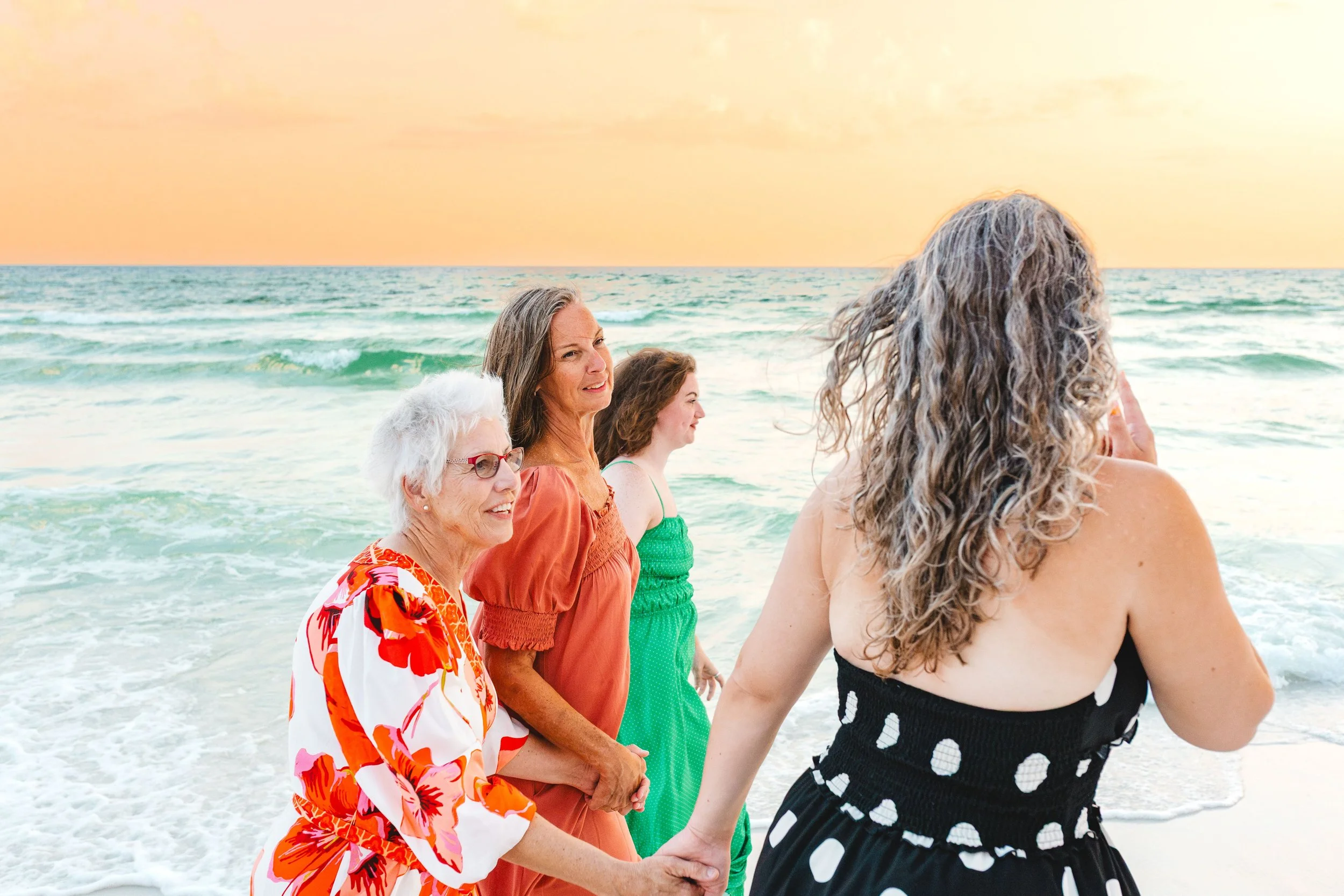 A group of women holding hands and praying on the beach during sunset.