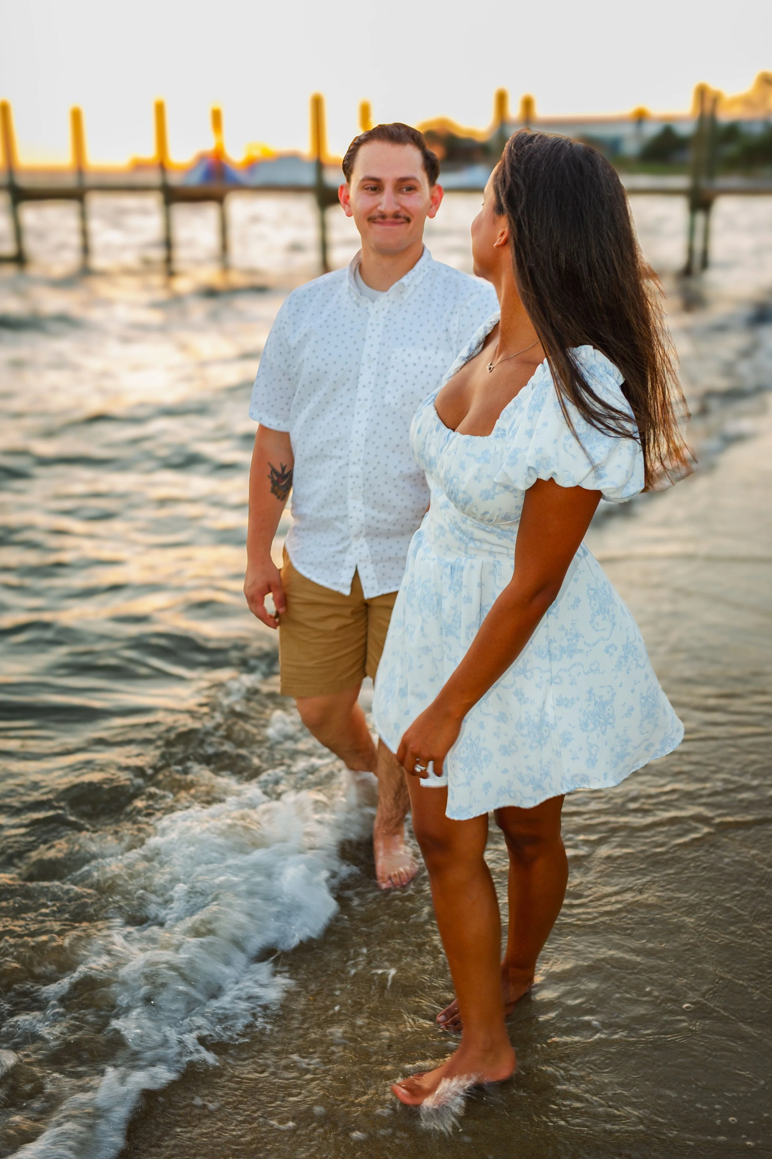 A couple standing in shallow water at a beach during sunset, facing each other and smiling, with a pier and boats in the background.