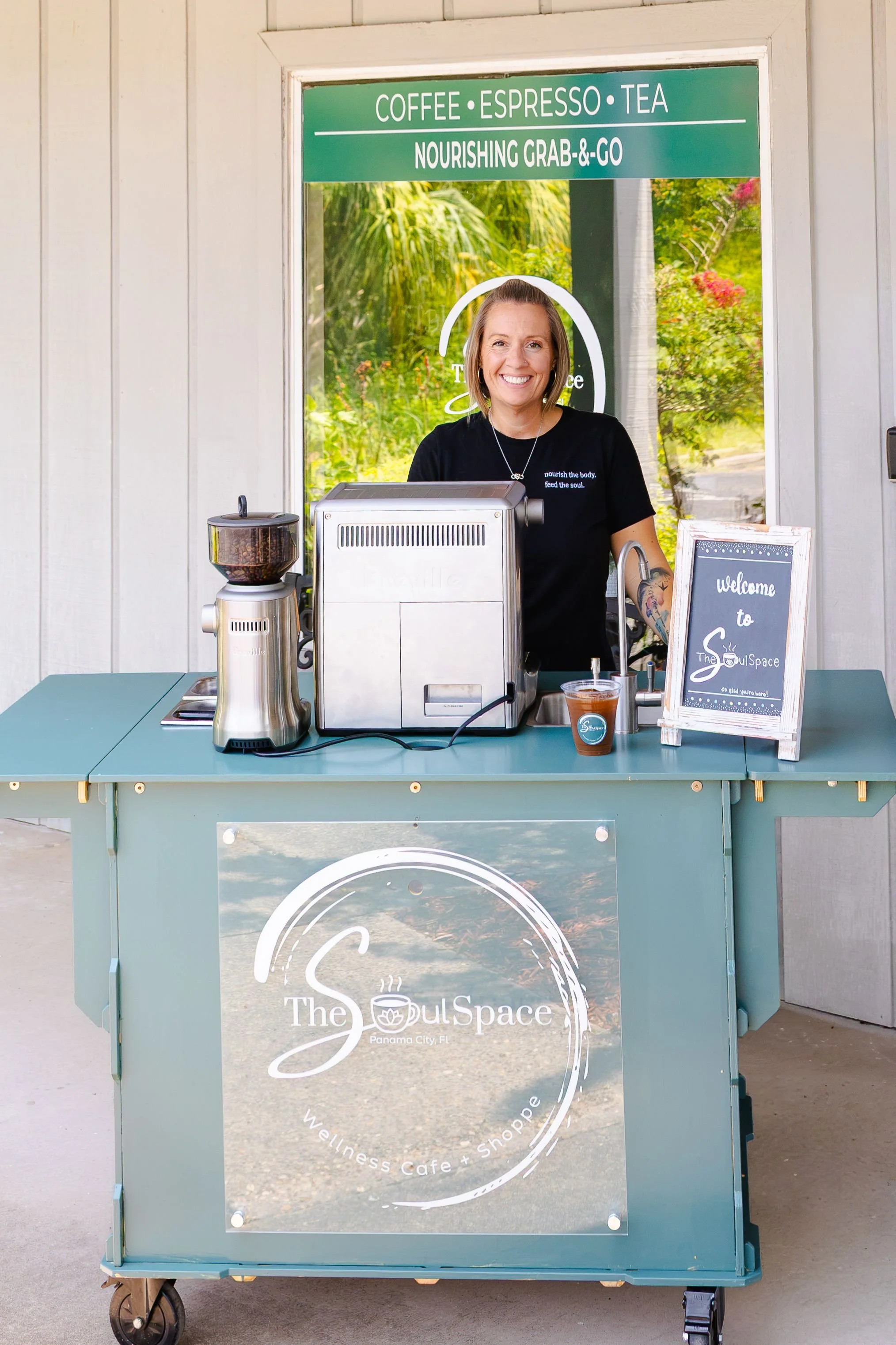 A woman standing behind a teal selling cart with a logo that reads "The Soul Space" and "Wellness Cafe + Shoppe" at Panama City, FL. The cart has coffee equipment and a small chalkboard sign that says "welcome to The Soul Space". She is indoors with 
