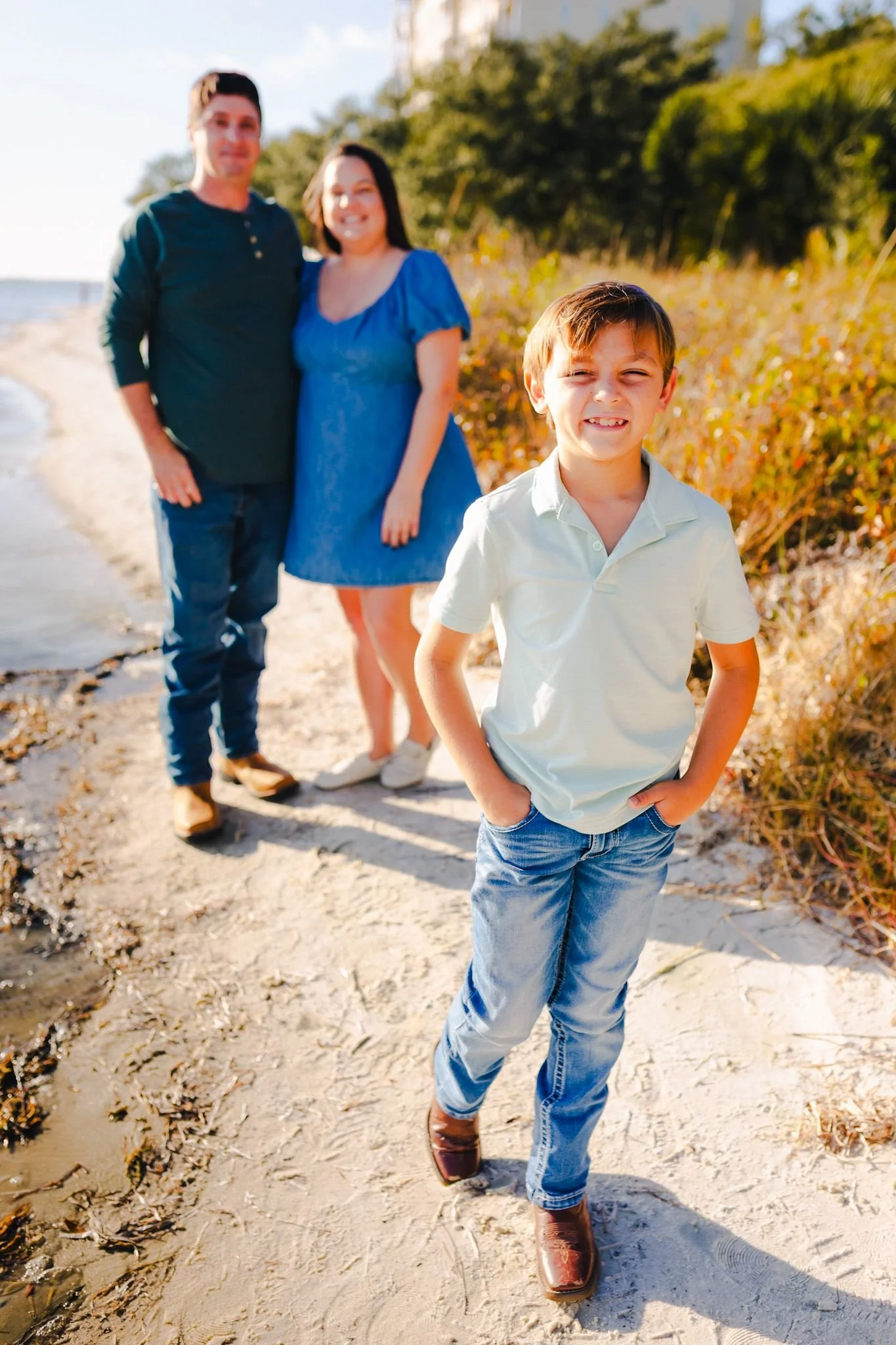 A family of three, consisting of a man, woman, and young boy, walking along a sandy beach with trees in the background during the daytime.