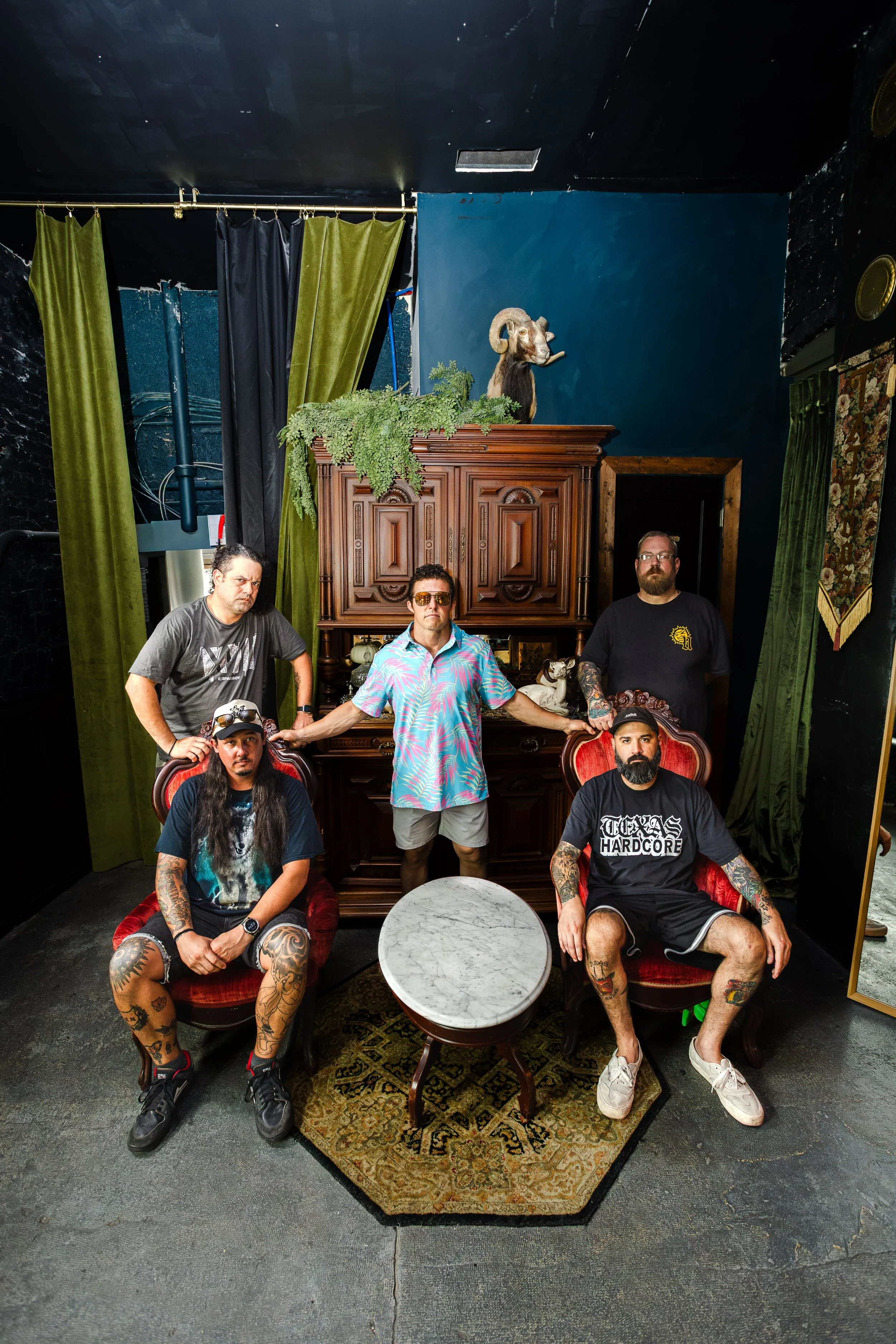A group of six men with tattoos and casual clothing posing in a room with vintage furniture, green and black curtains, and decorative elements. There is a wooden cabinet with a mounted ram's head and greenery on top. The men are sitting and standing 