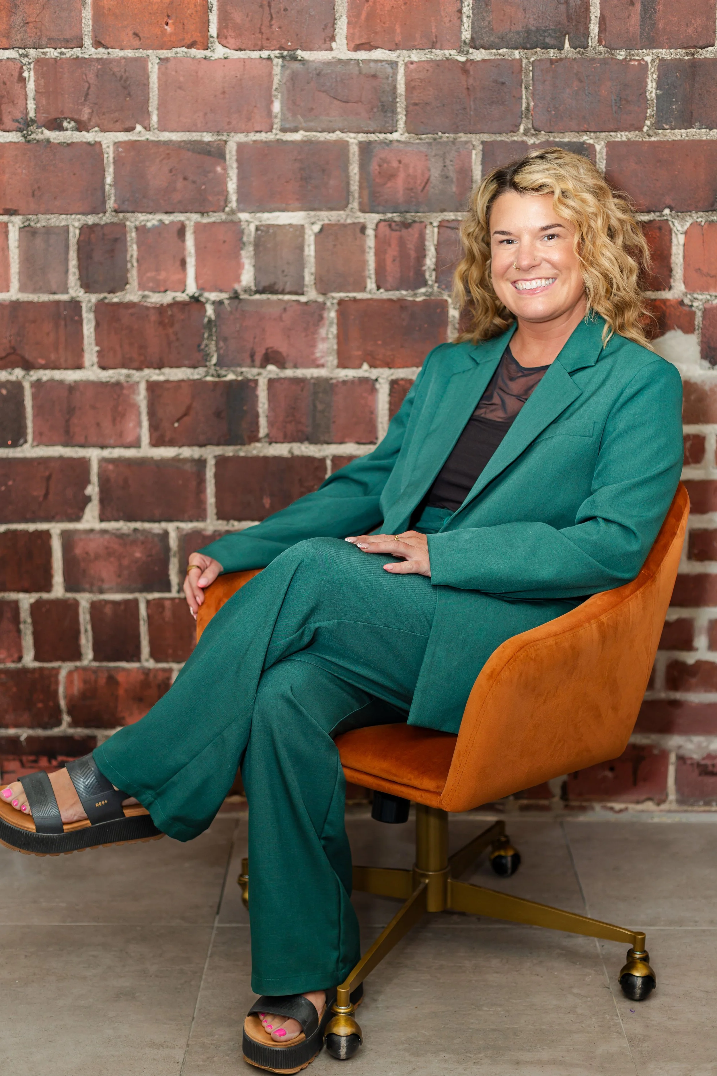 A woman with curly blonde hair sitting in an orange chair in front of a brick wall. She is smiling and wearing a teal suit with a black top and black sandals.