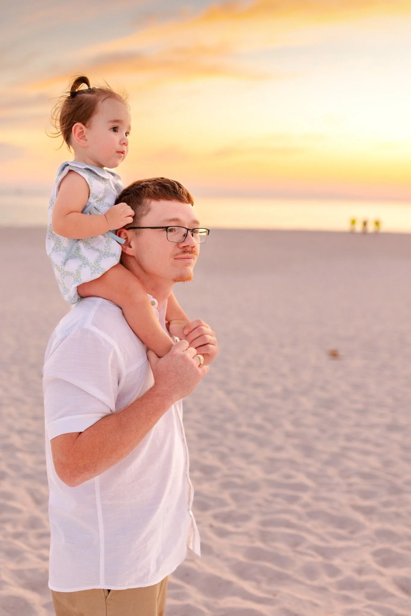 A man with glasses and a short-sleeved white shirt carrying a young girl on his shoulders at the beach during sunset. The sky is colorful with orange, pink, and yellow hues, and the beach appears peaceful with a few people visible in the distance.