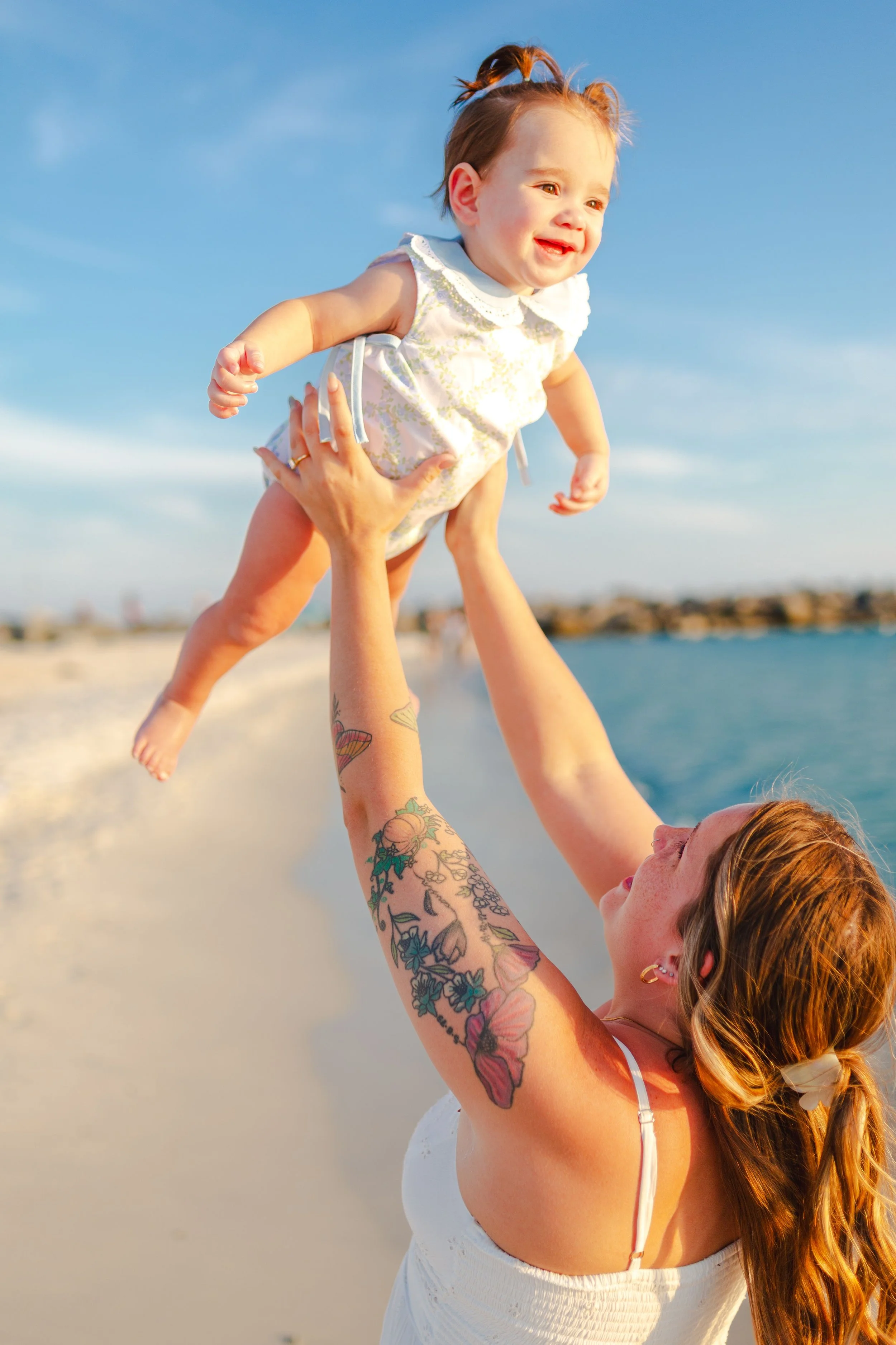 A woman with red hair and tattoos lifting a young girl with light hair and a white dress at the beach during sunset, with a blue sky and ocean in the background.