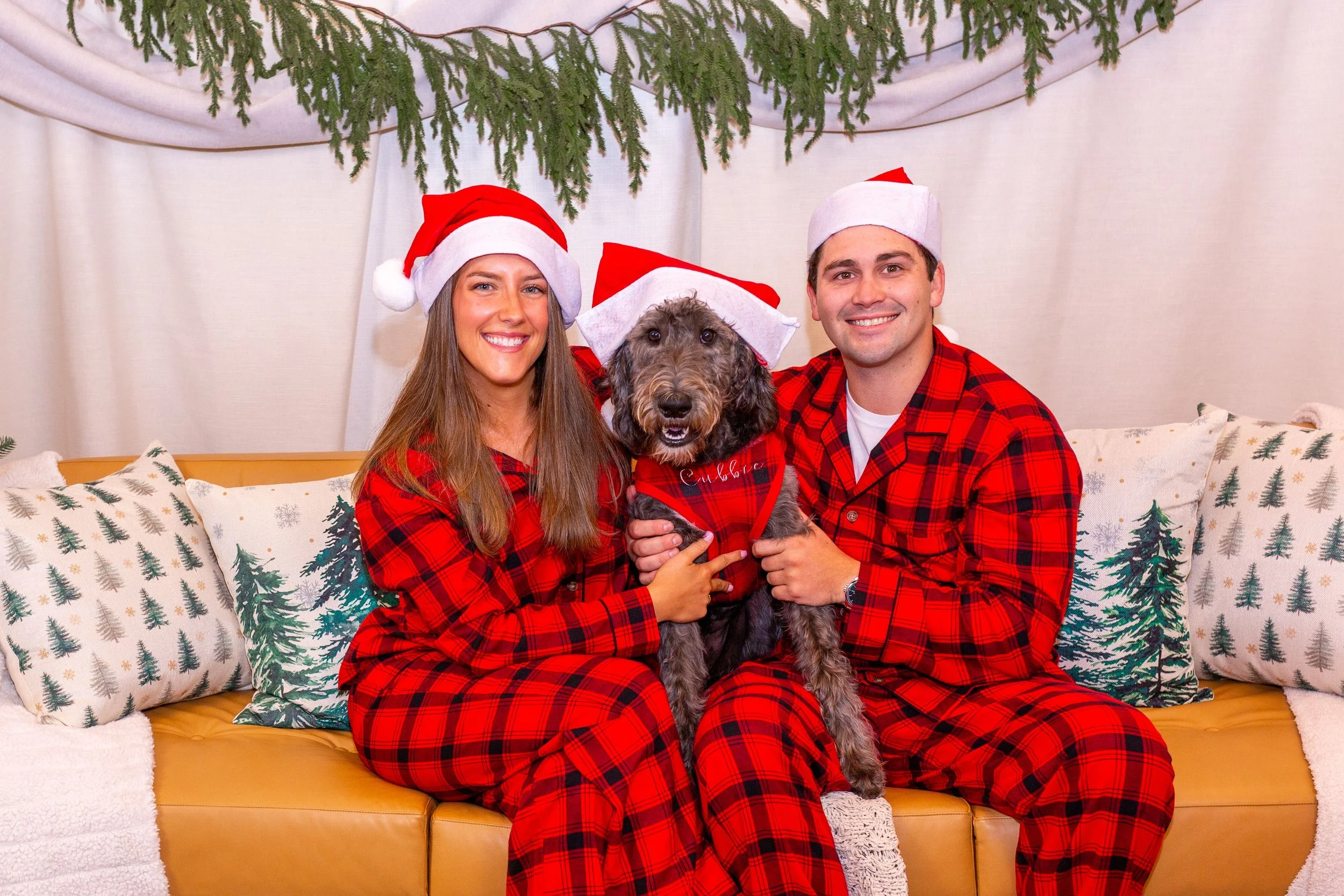A smiling man and woman dressed in red Christmas pajamas and Santa hats sitting on a sofa, holding a dog wearing a holiday bandana. They have a festive backdrop with Christmas greenery and decorative pillows with Christmas trees.