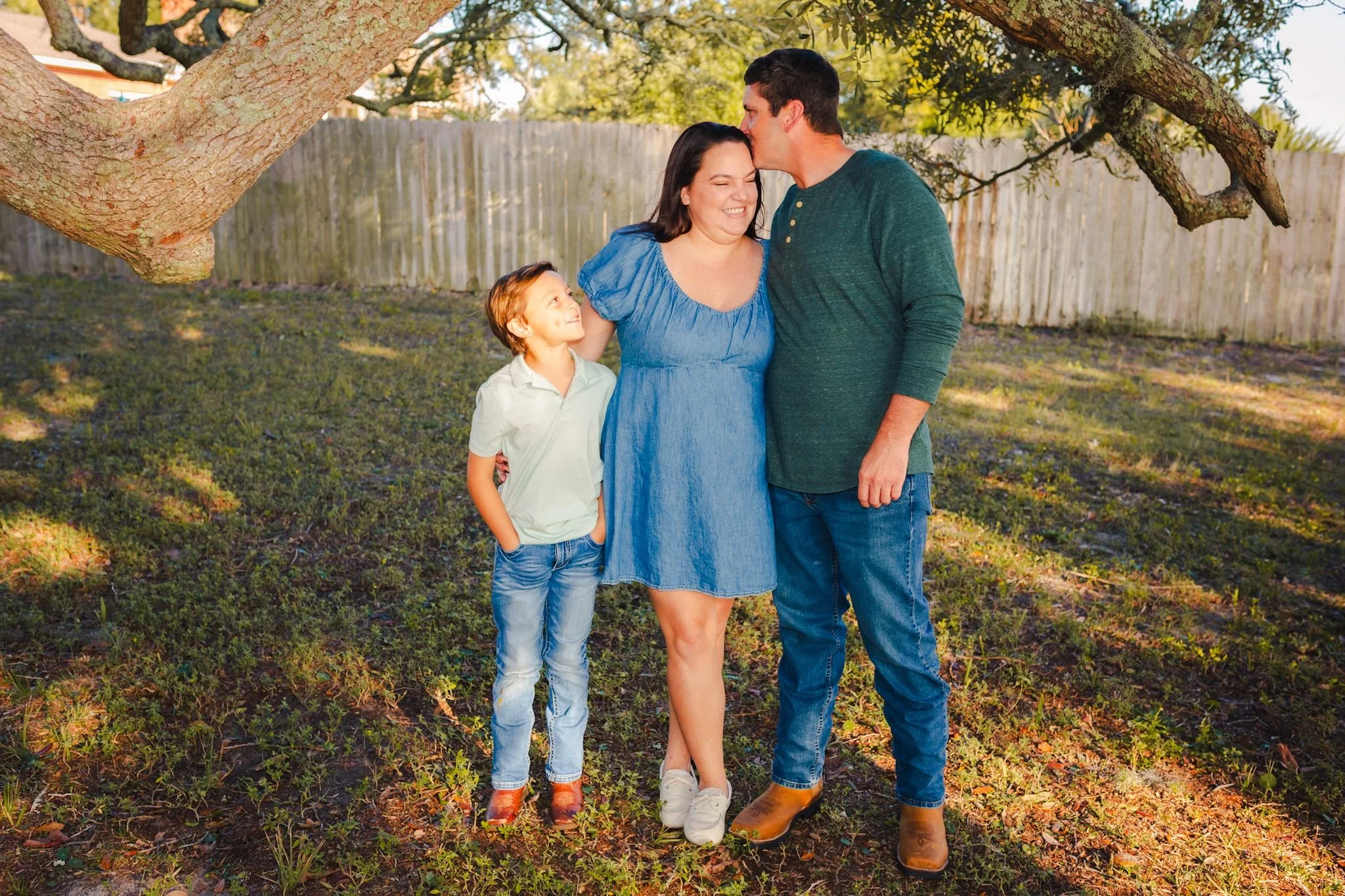 A family of three standing outdoors near a large tree, with the father kissing the mother's forehead, the mother smiling, and the young son looking up at her, all enjoying a sunny day in a backyard.