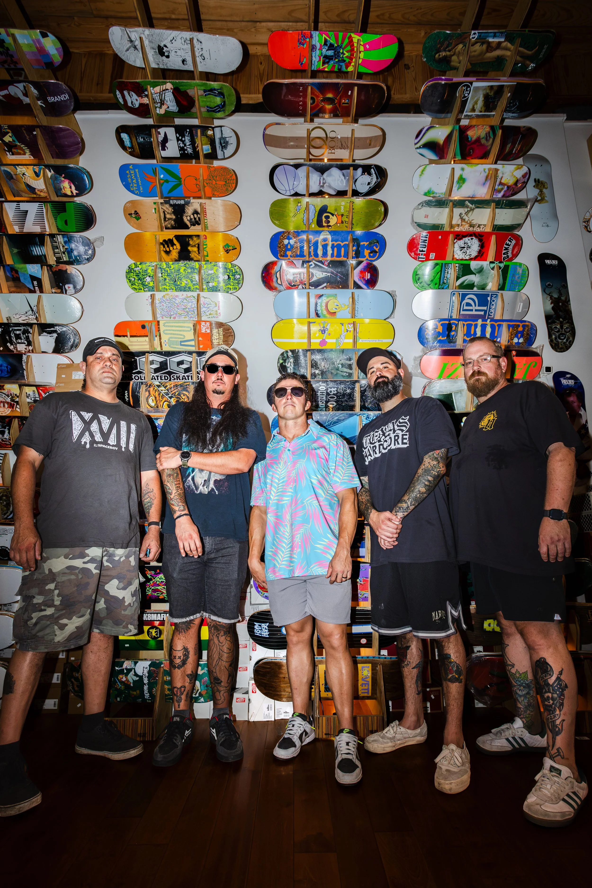 Five men standing in front of a wall display of skateboards, with various skateboard designs and artwork.