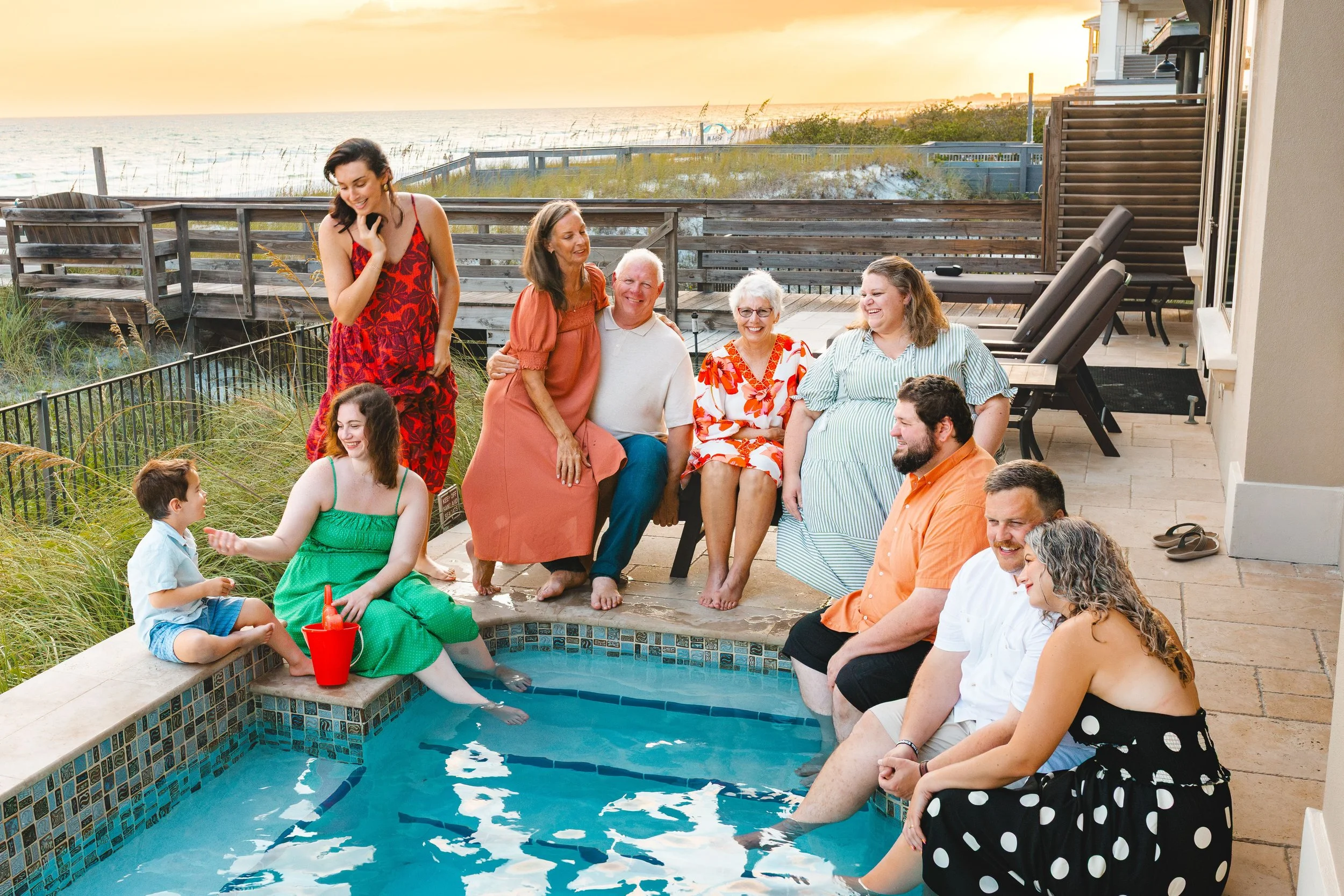 A group of people gathered on a patio near a swimming pool with a view of the beach and ocean during sunset. Some are sitting on the edge of the pool with their feet in the water, while others stand and sit nearby, smiling and enjoying the moment.
