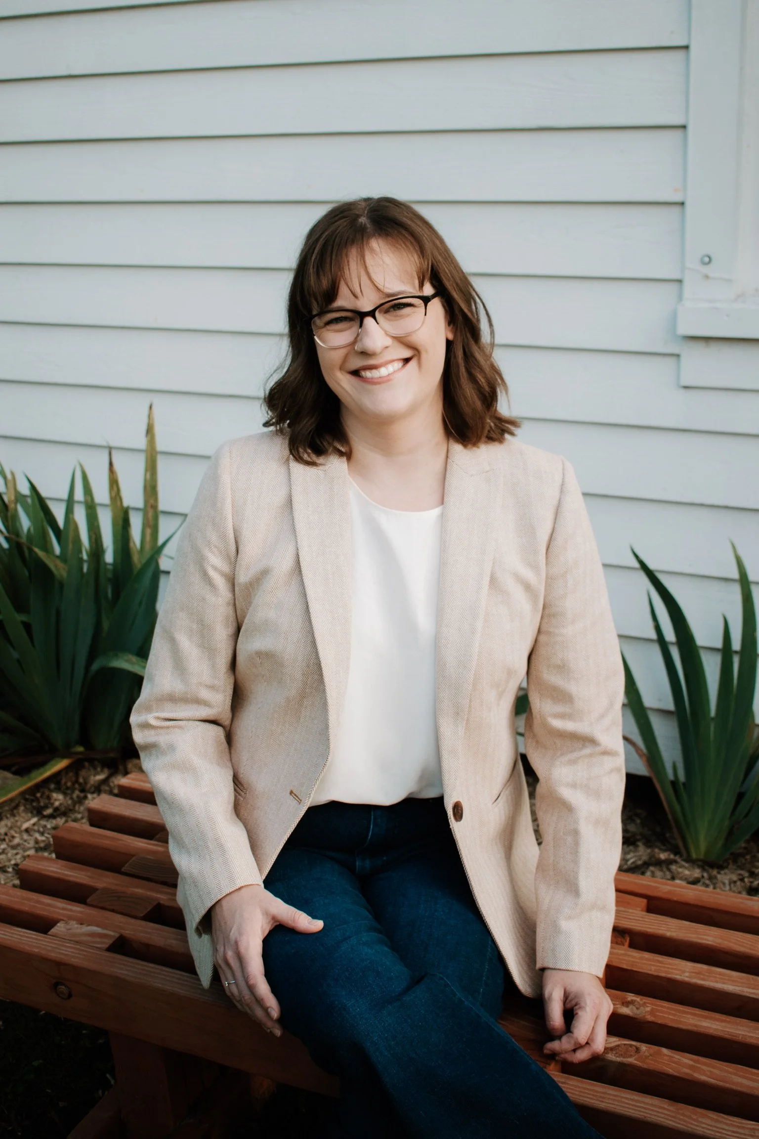 A woman with shoulder-length brown hair, glasses, and a warm smile, wearing a beige blazer and white blouse, sitting on a wooden bench outside with green plants and a white house siding background.