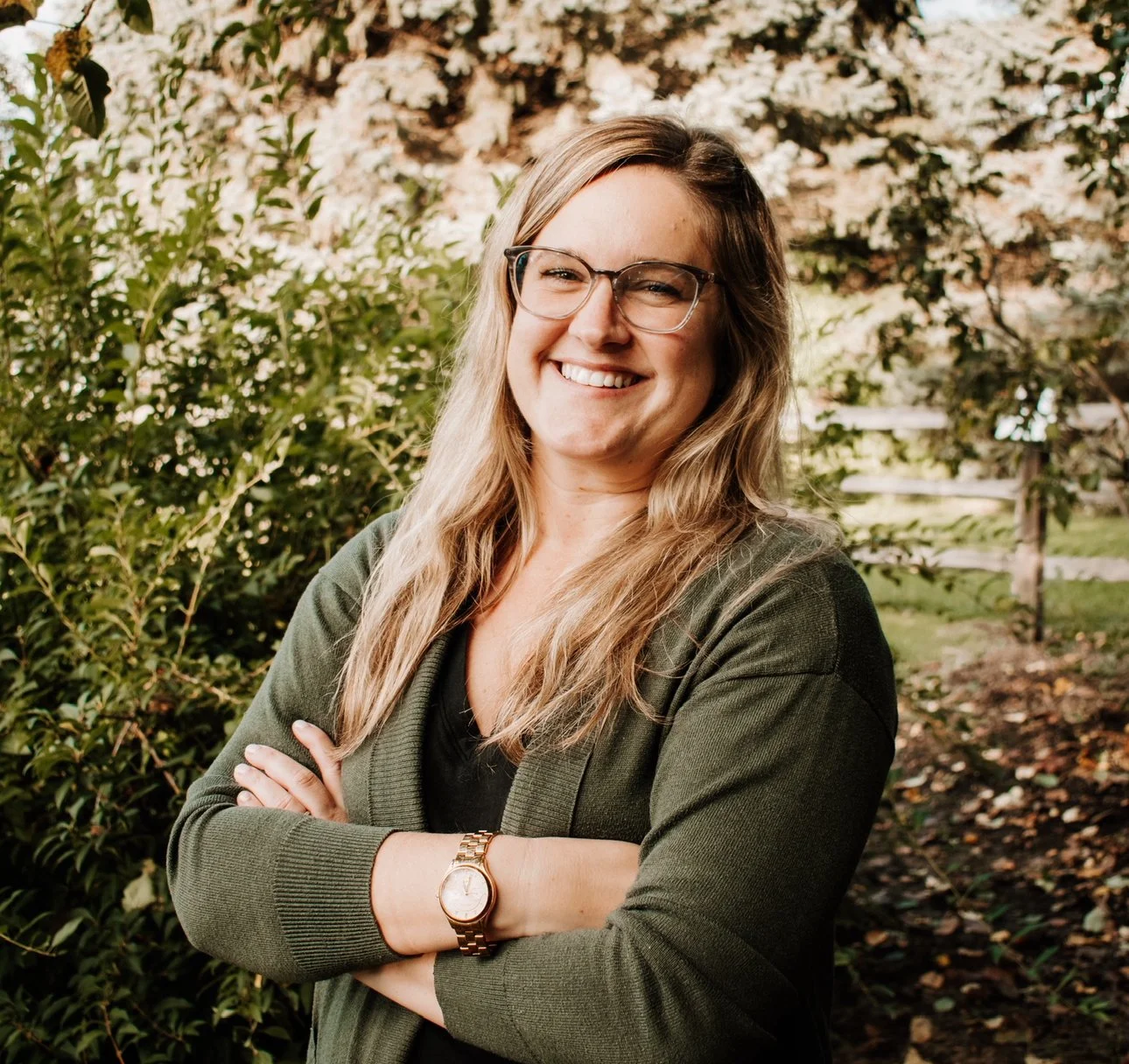 A woman with long blonde hair, glasses, and a warm smile stands outdoors with her arms crossed, surrounded by greenery and trees in the background.