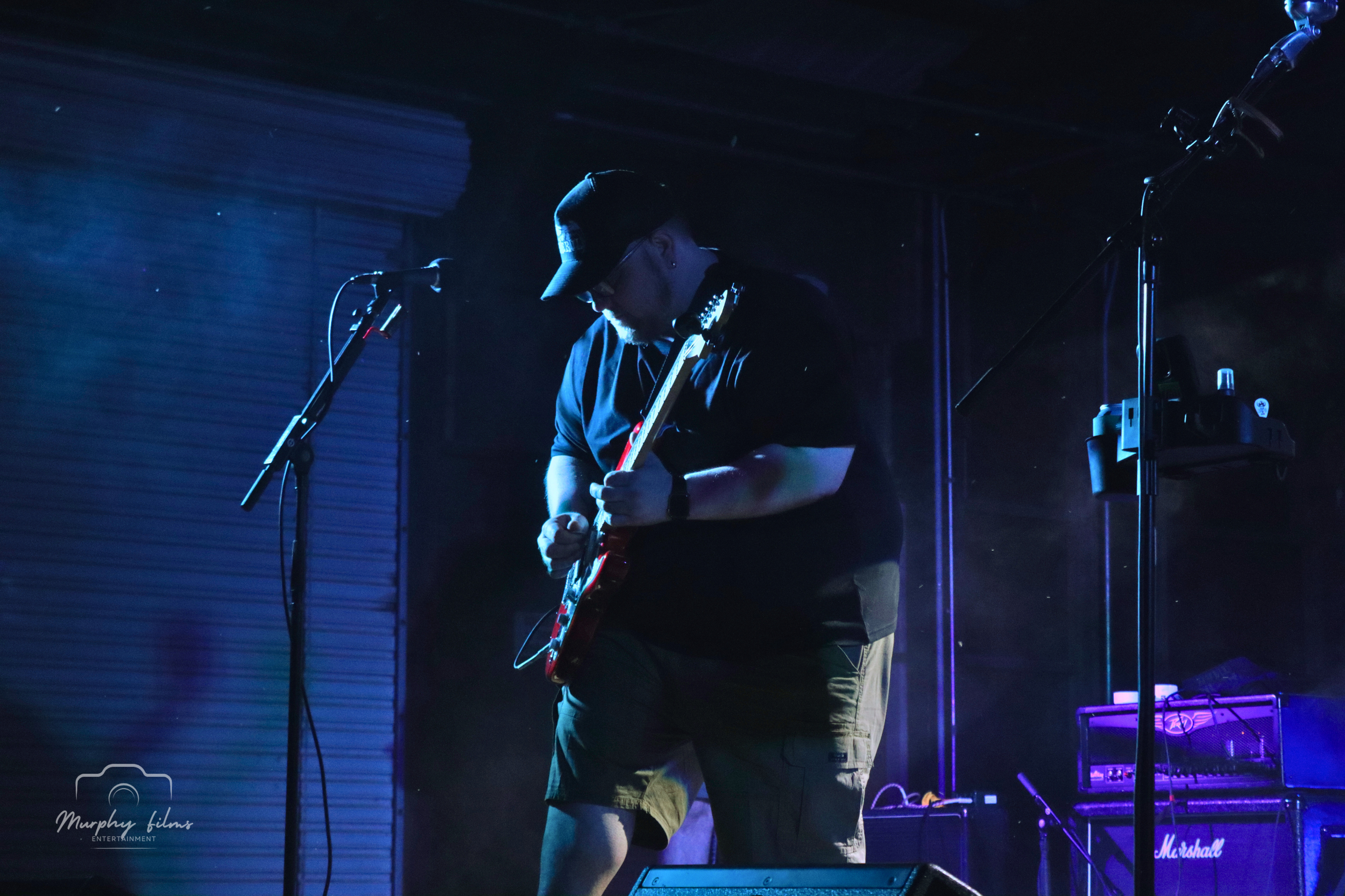 Musician playing electric guitar on stage with microphones and amplifier, dim blue and purple lighting.