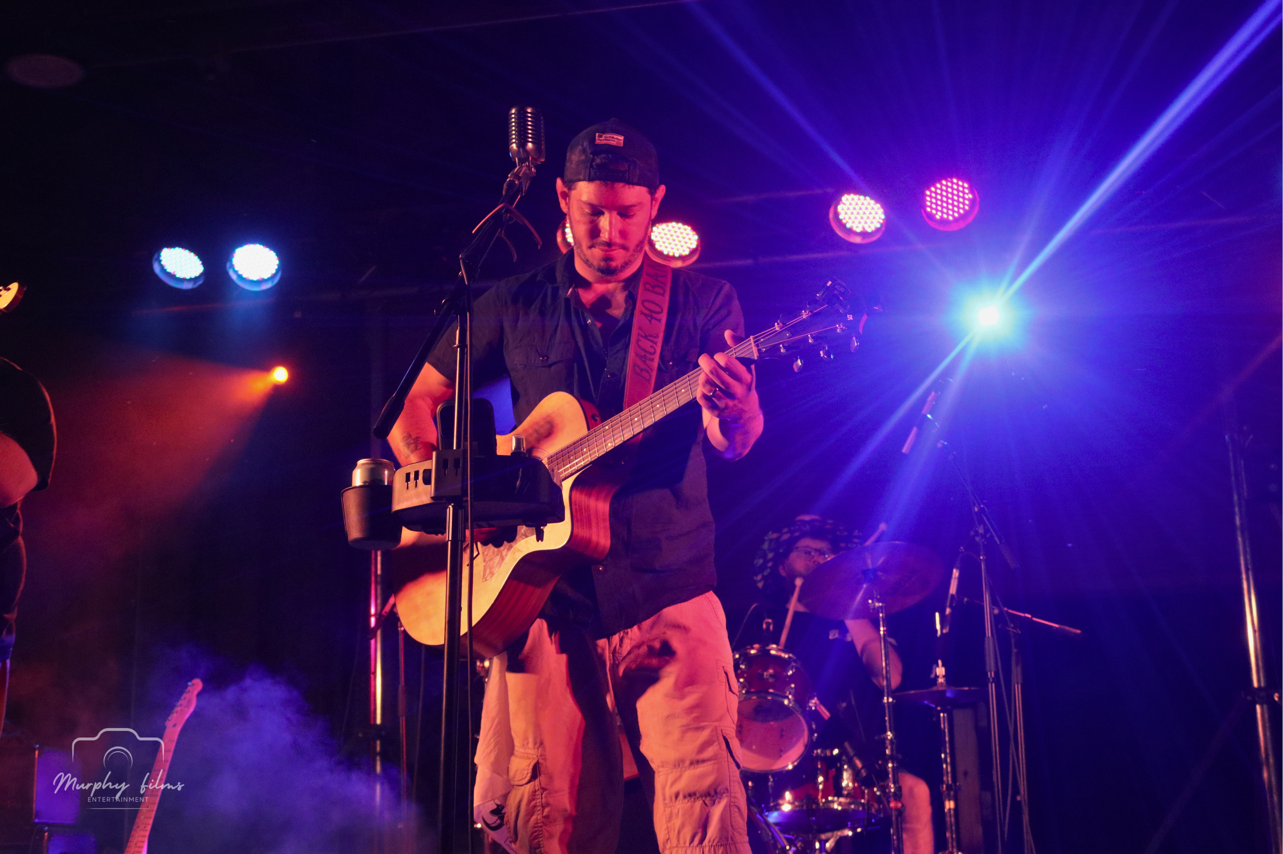 Musician playing an acoustic guitar on stage, illuminated by colorful purple and yellow stage lights, with a drummer in the background.