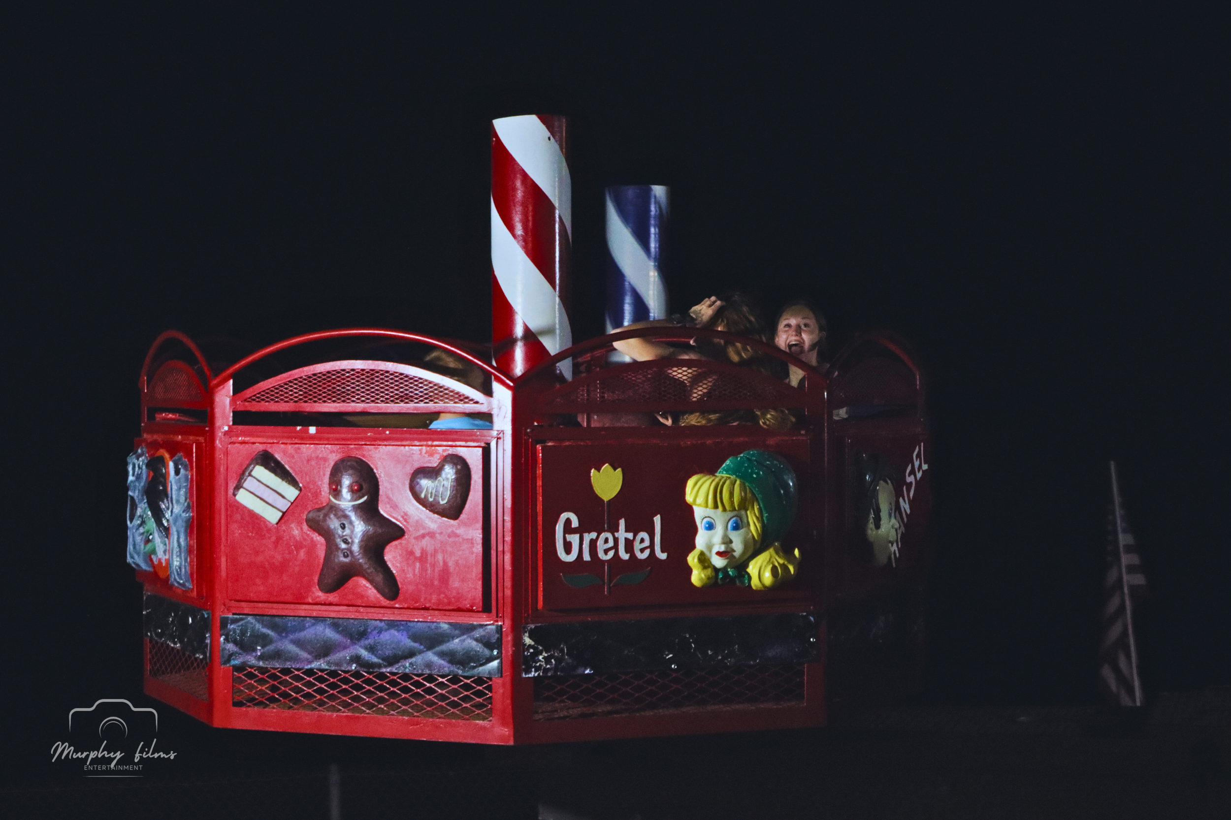 A red carnival ride featuring the character Gretel from the Hansel and Gretel fairy tale, decorated with colorful candies and gingerbread men, with two children enjoying the ride at night.