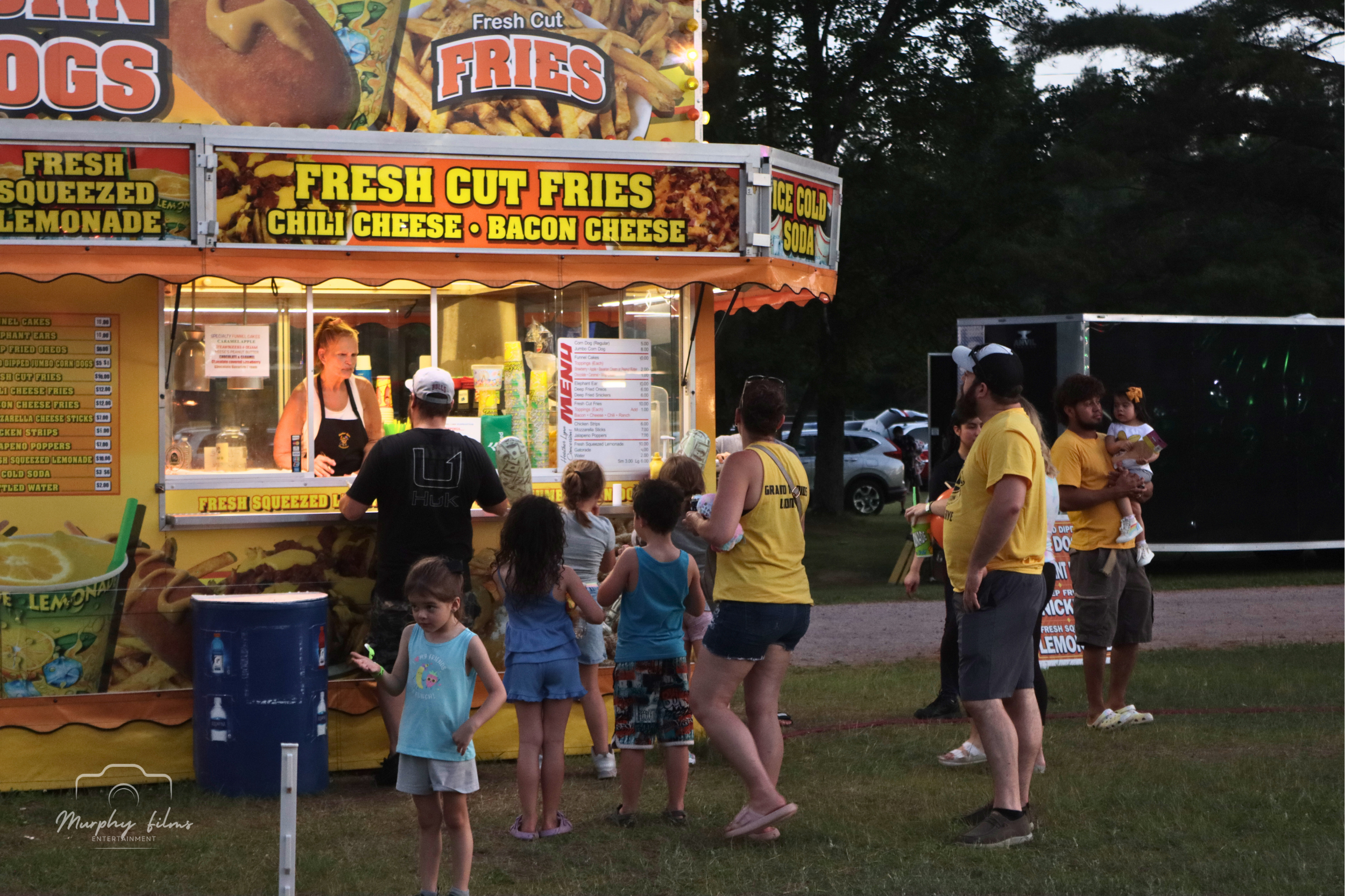 At a carnival food stand selling fresh cut fries with chili, cheese, and bacon cheese, several people, including children and adults, are gathered in line and socializing. The stand features vibrant signs and a menu, with a line of cars and trees in