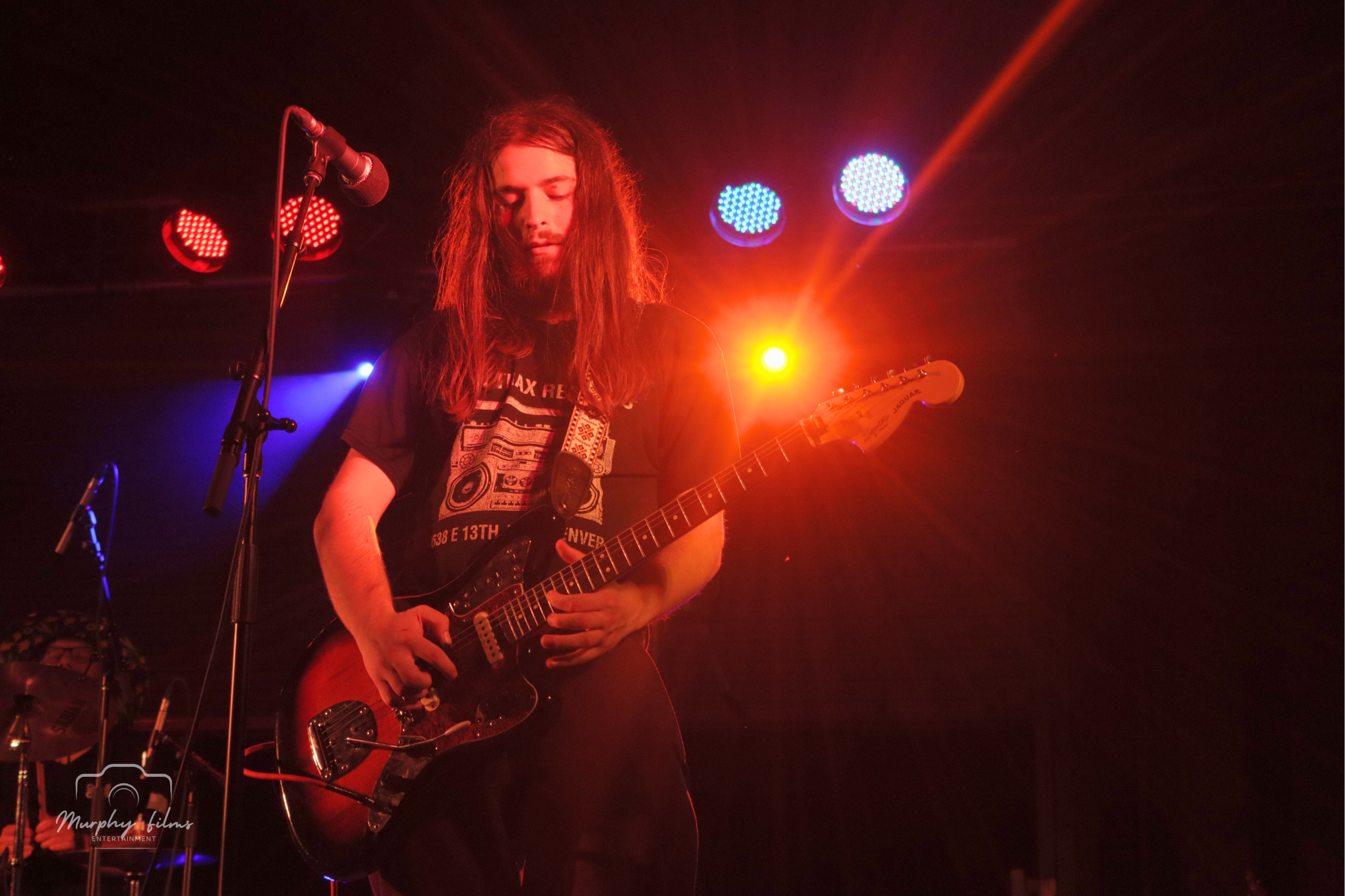 A young male guitarist with long hair and a beard performing on stage under vibrant, colorful stage lights.