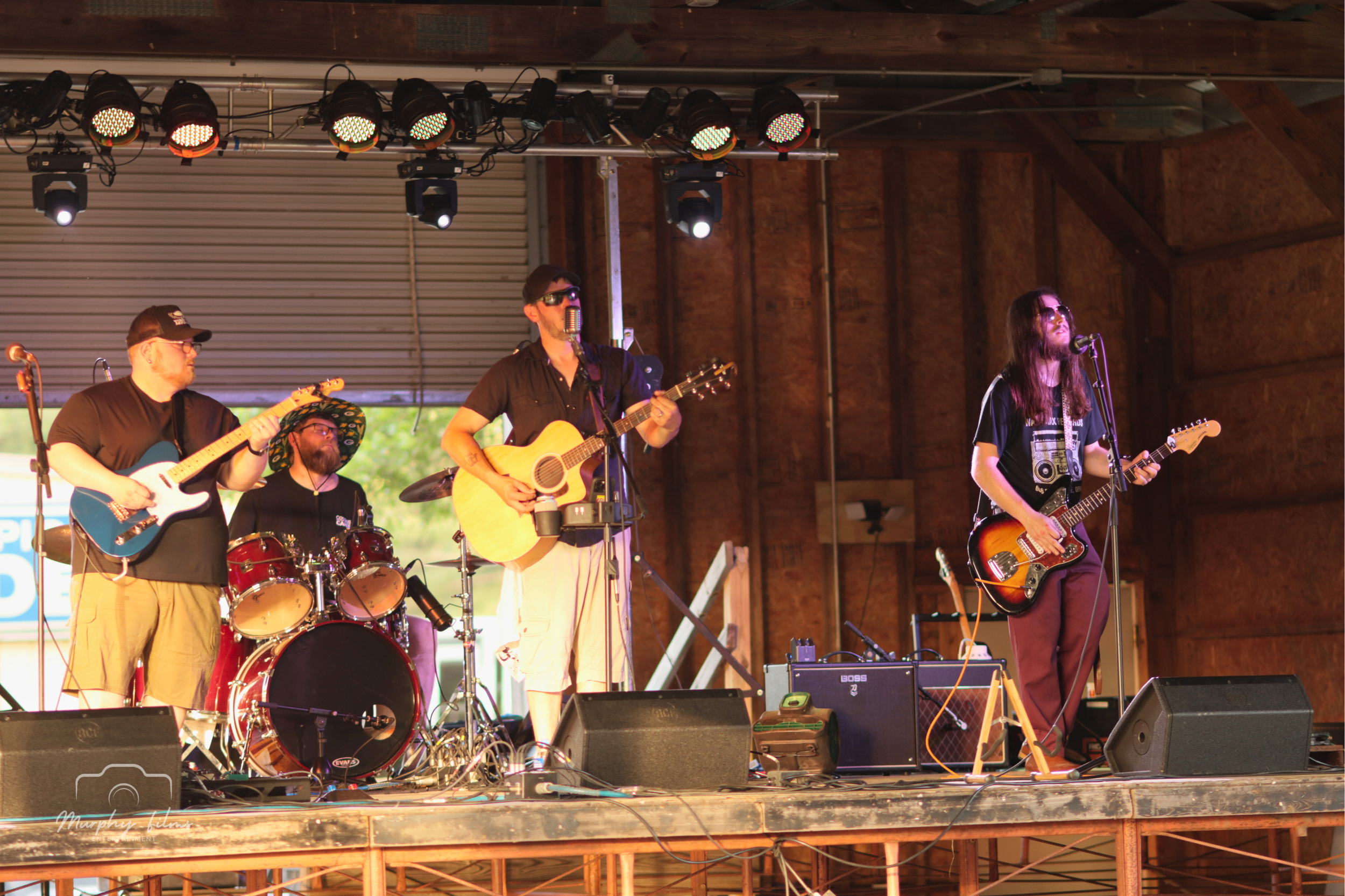 A band performing on a stage with four members playing guitars and drums, inside a wooden structure with stage lights overhead.