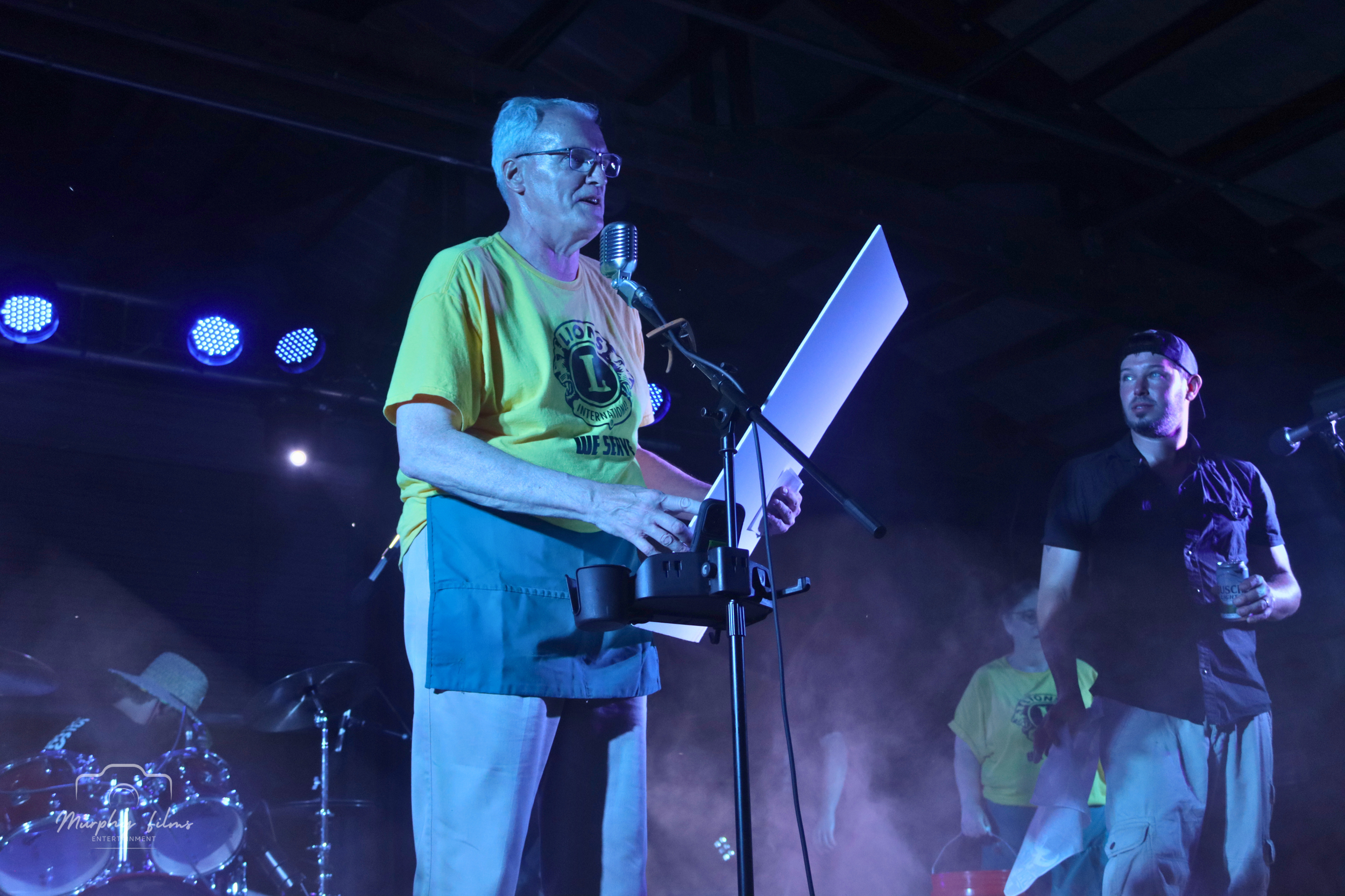 An older man with glasses standing on stage, reading from a paper into a microphone, while another man holds a drink nearby. The stage is lit with blue and purple lights, with musical instruments visible in the background.