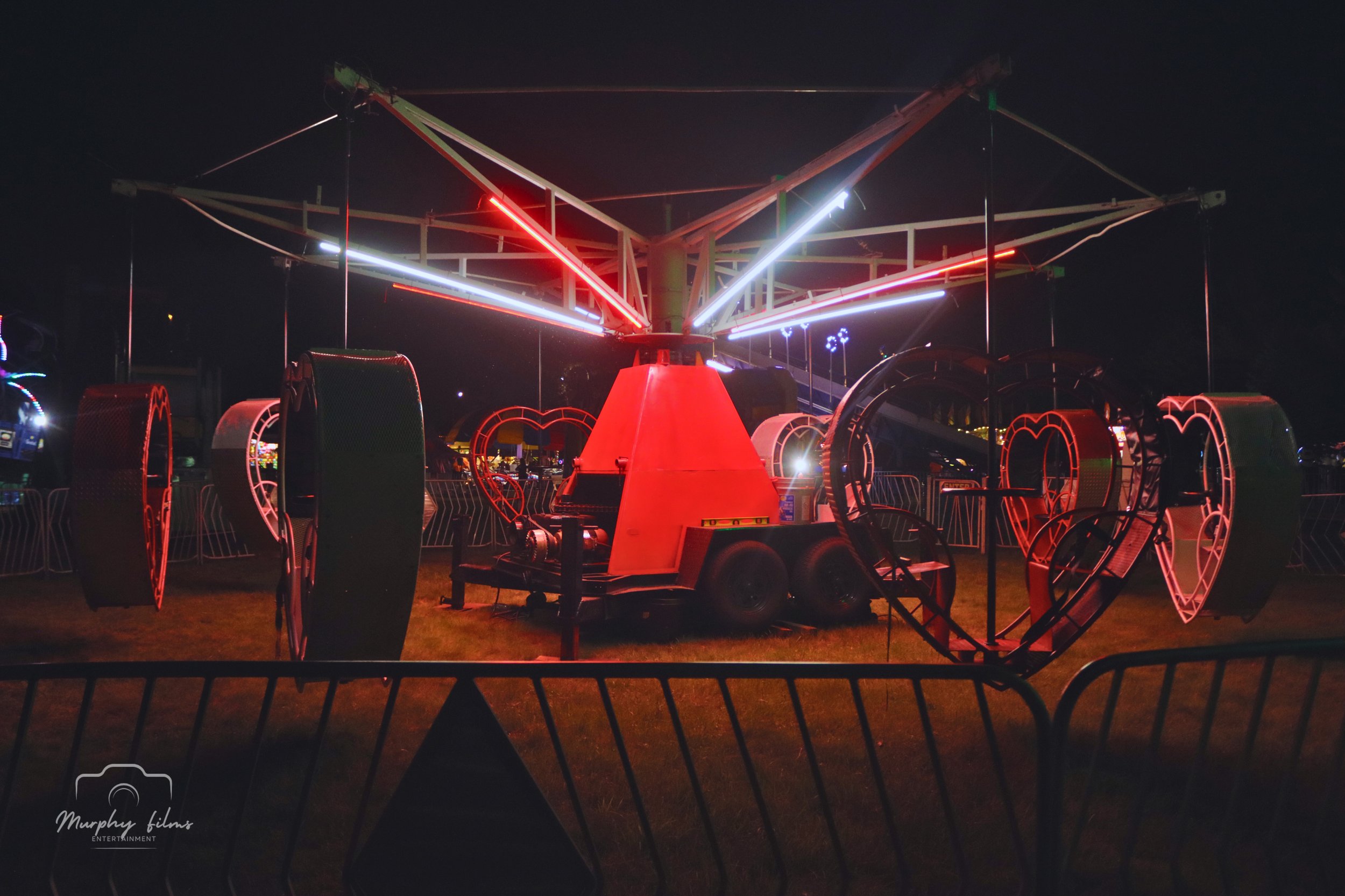 Lighted amusement ride at night with neon lights, heart-shaped elements, and a large rotating structure surrounded by safety barriers.
