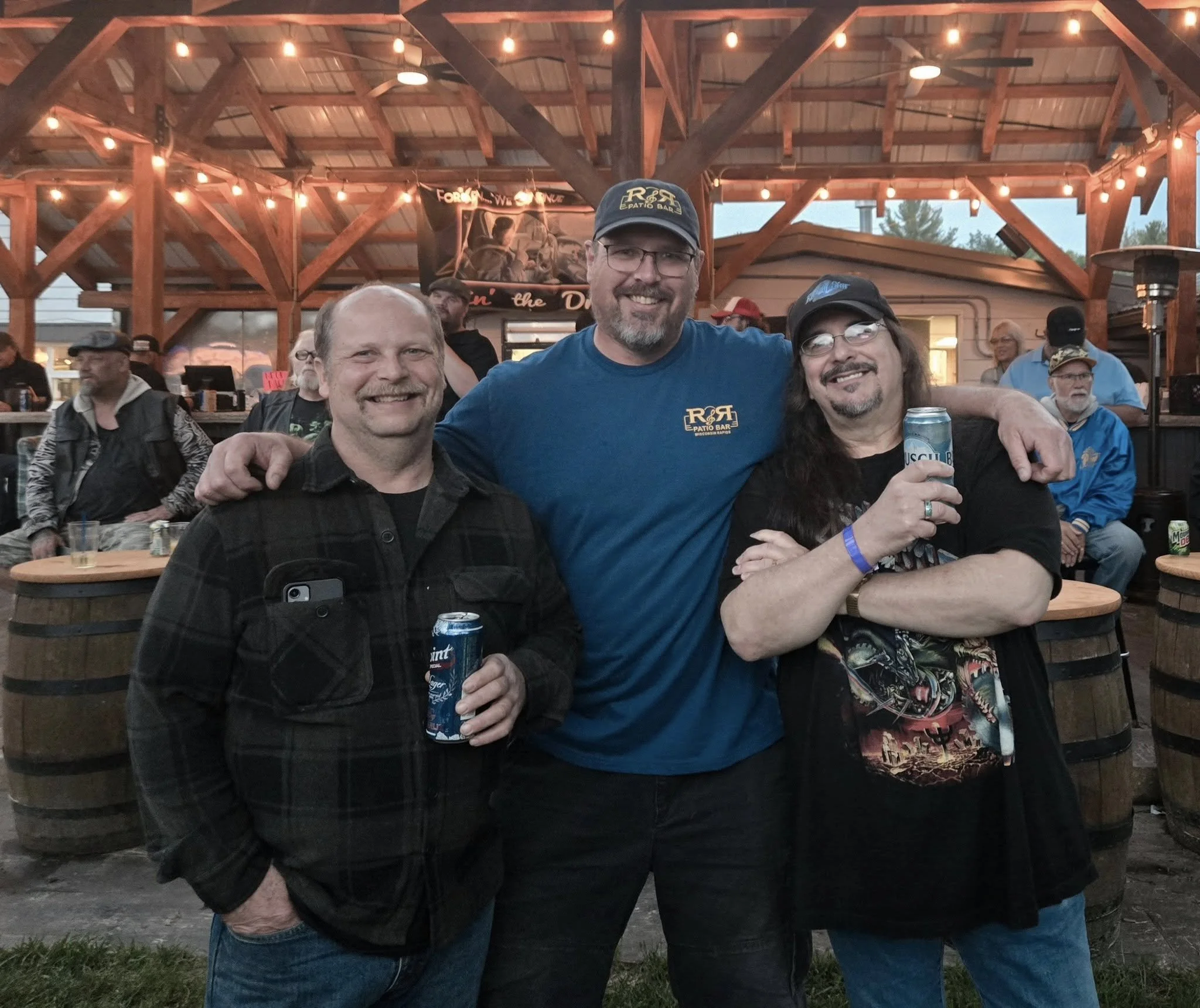 Three men smiling and holding drinks pose together at an outdoor bar with a wooden pavilion and string lights, with other people seated in the background.