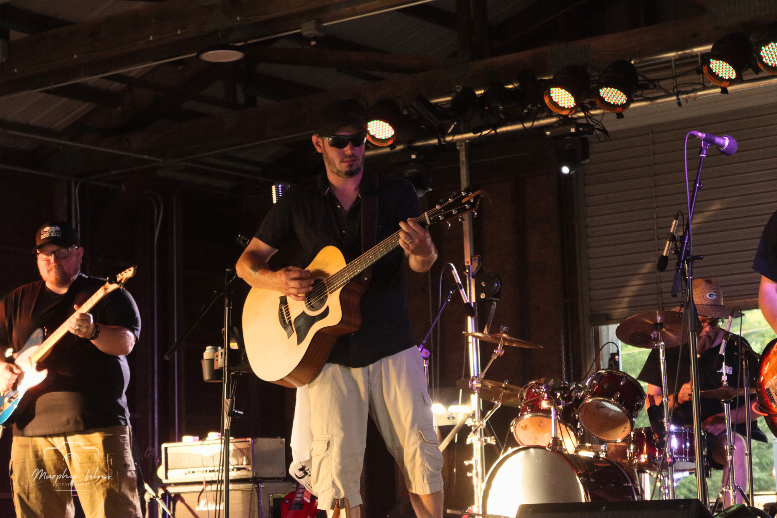 Musicians playing electric guitar, acoustic guitar, and drums on stage during a live performance, with stage lights overhead.