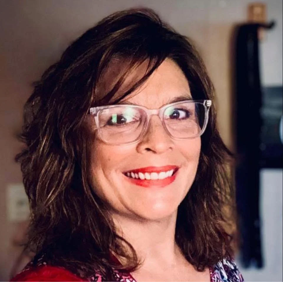 A woman with wavy brunette hair, wearing clear glasses, red lipstick, and a red and black patterned top, smiling at the camera in an indoor setting.