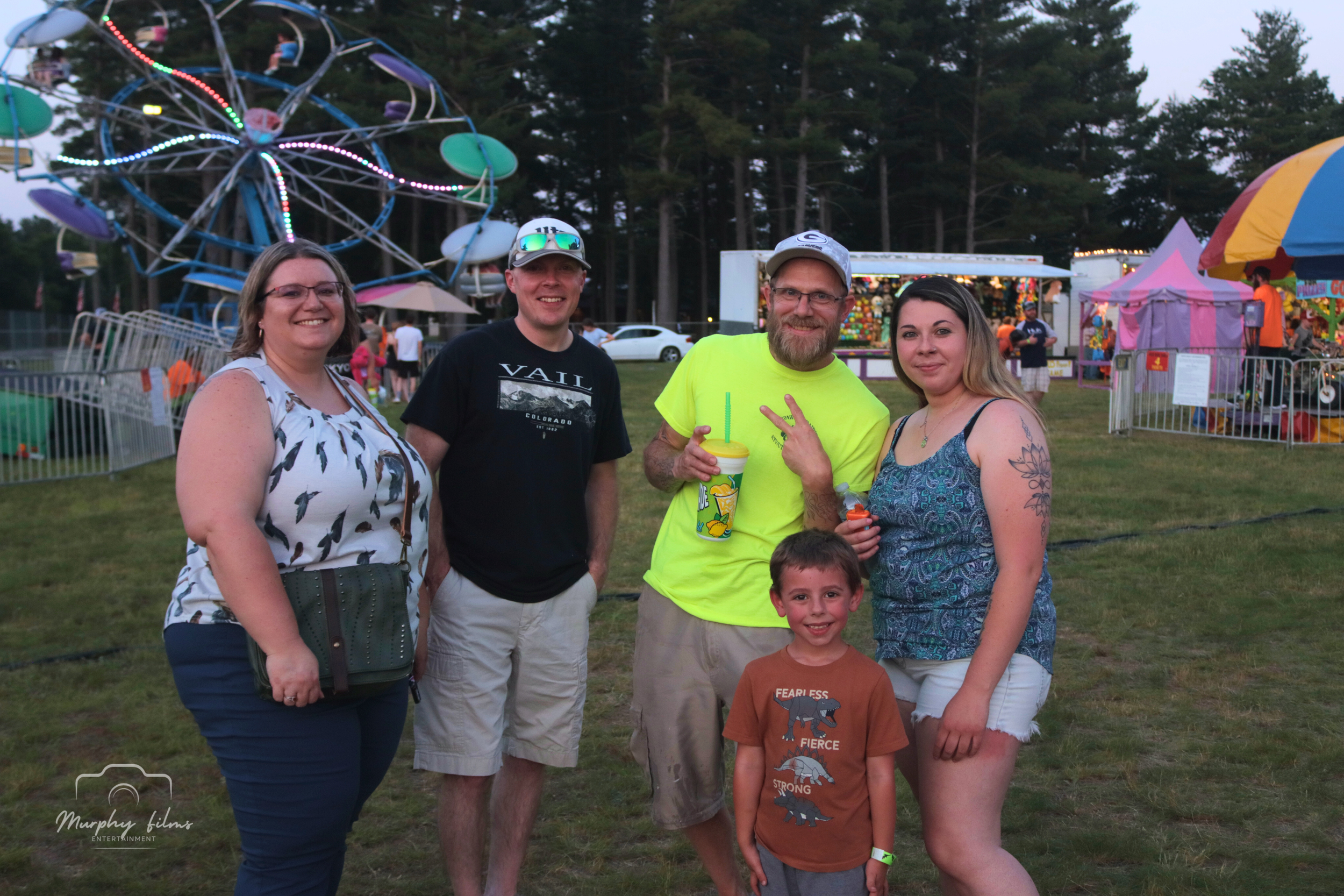 Group of five people, including two women, two men, and a young boy, standing together at a fairground with a Ferris wheel, amusement rides, and booths in the background