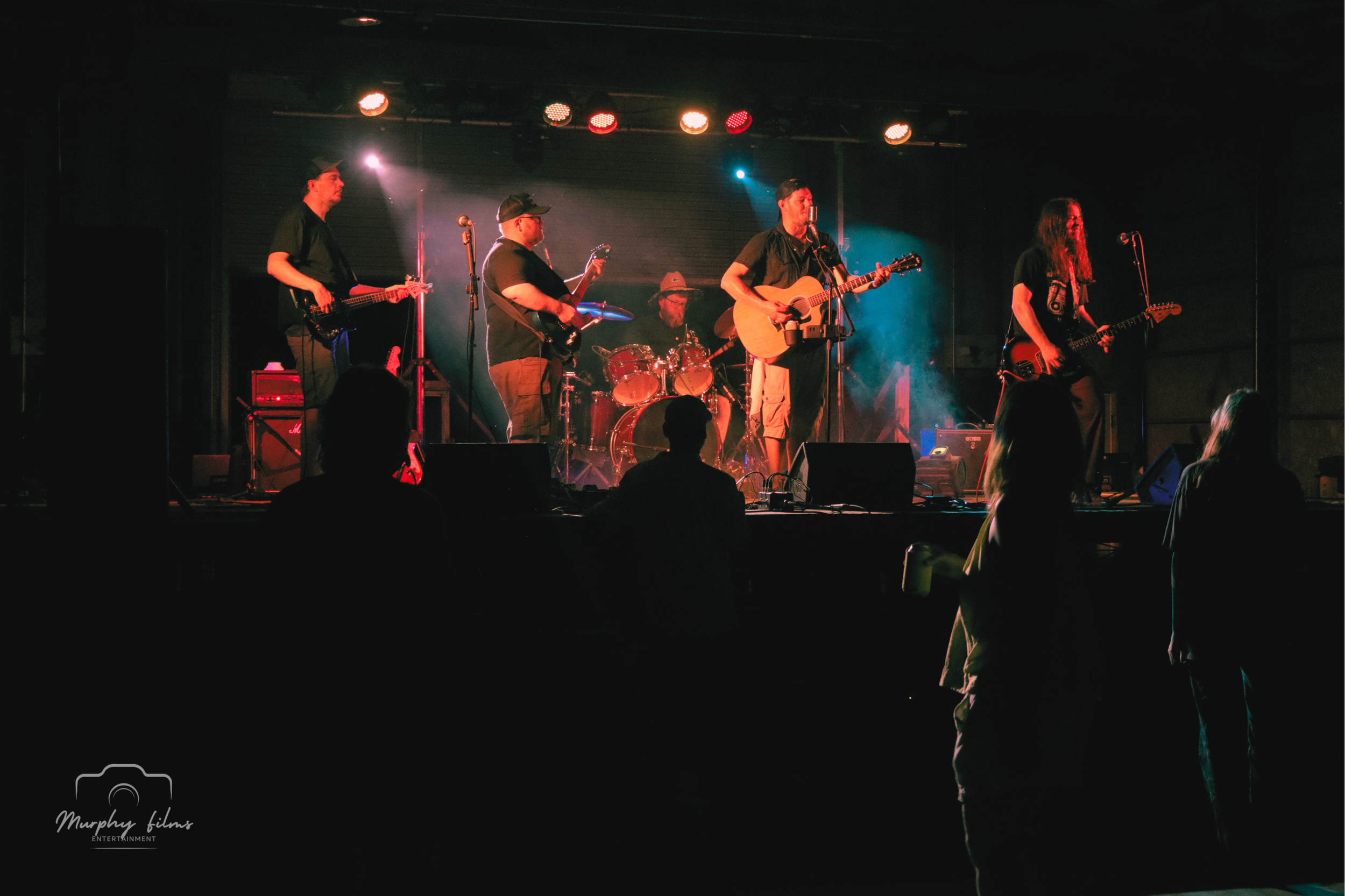 A live band performing on stage with five members playing guitars, drums, and vocals, illuminated by stage lights, with an audience watching in front.