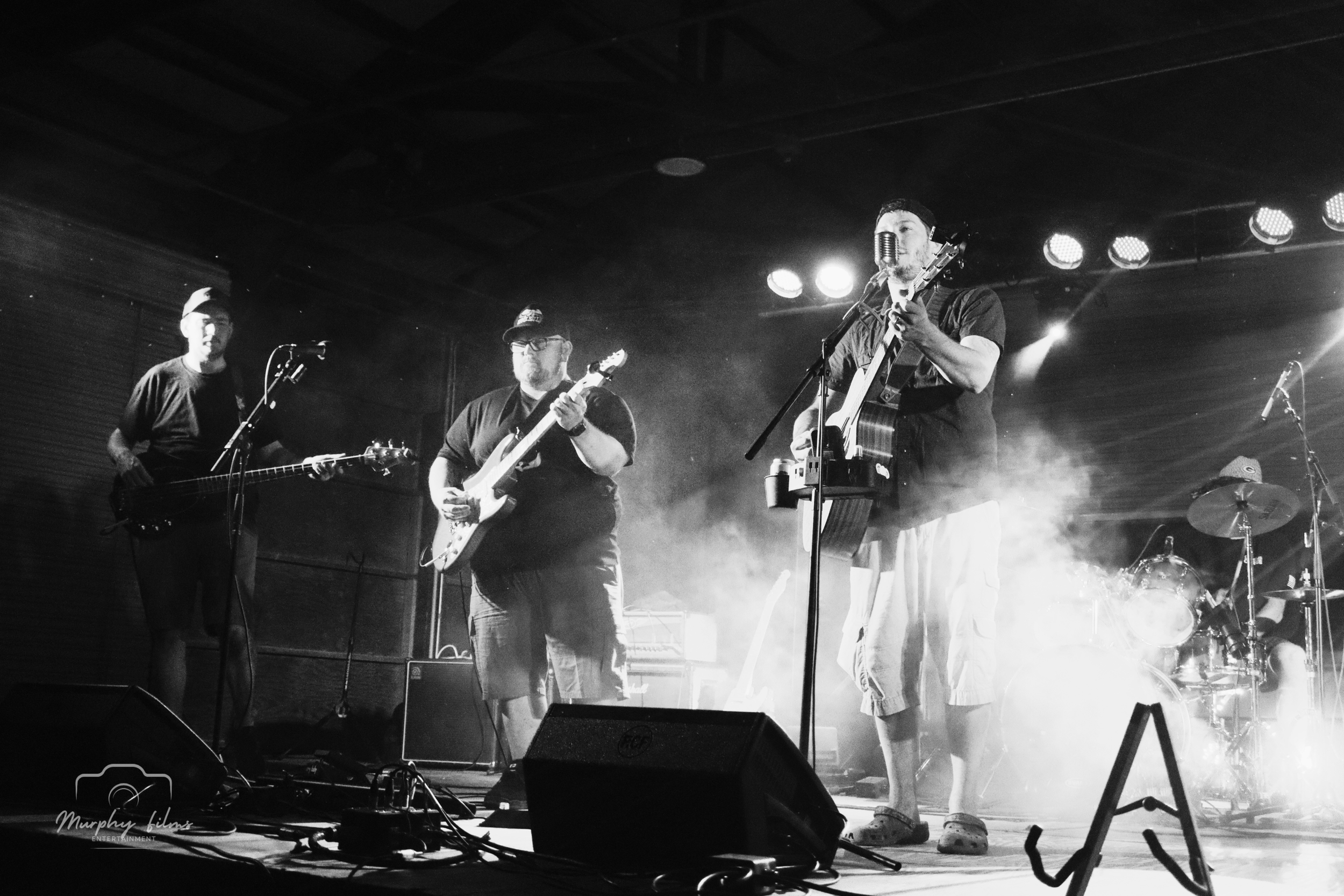 Black and white photo of a band performing on stage with three members playing guitars and one on drums, with stage lighting and fog effects.