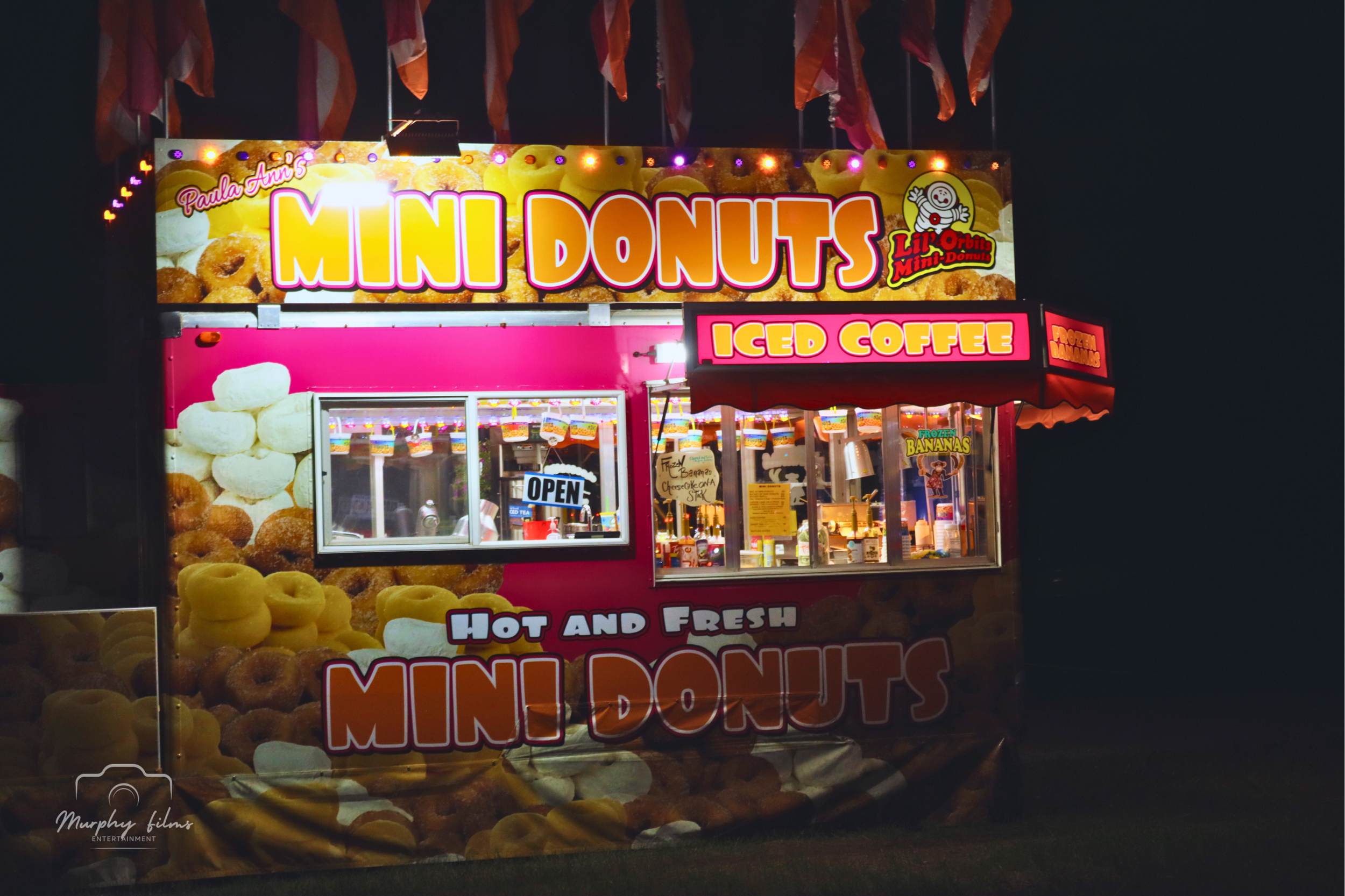 A brightly lit food stand offering mini donuts and iced coffee at night, with colorful signage and flags flying above.