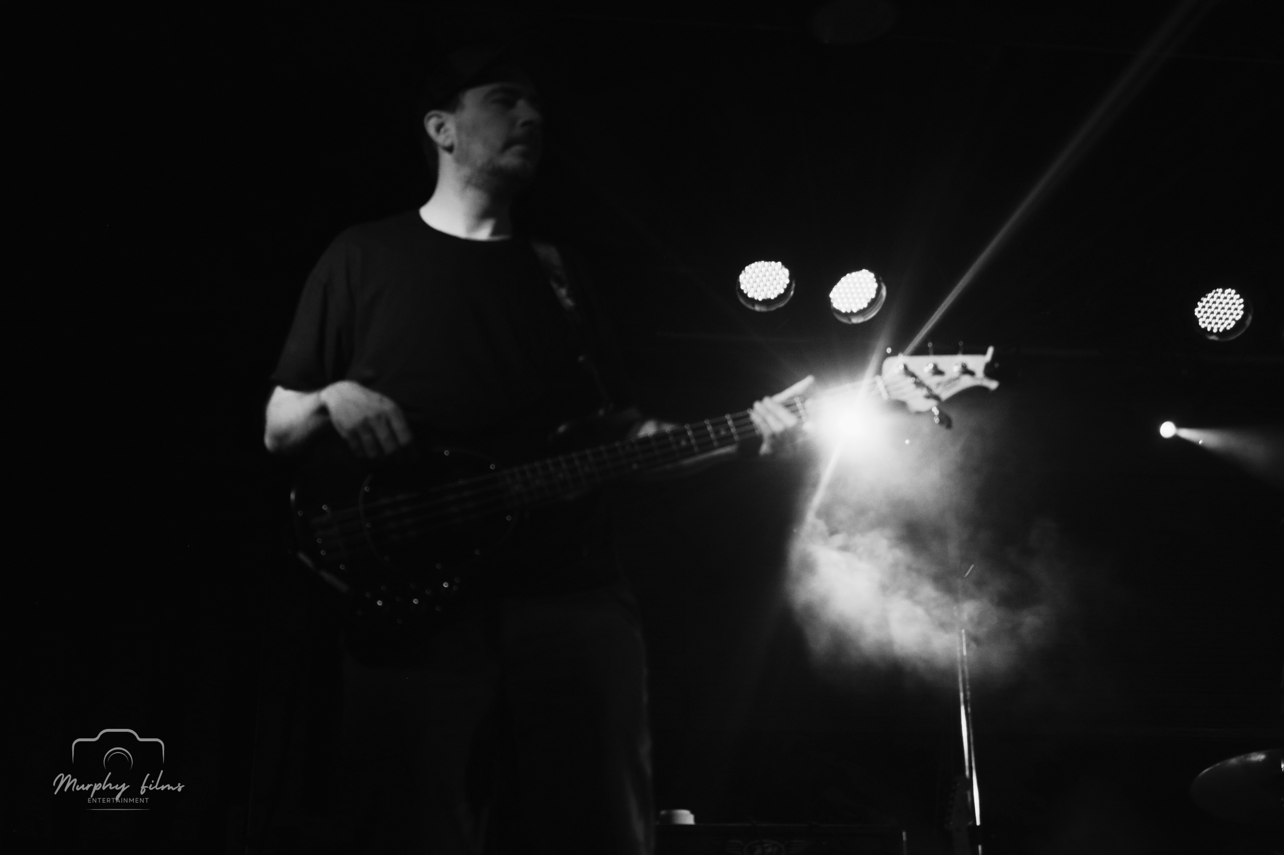 A person playing a bass guitar on stage, with bright stage lights and fog creating a dramatic atmosphere in black and white.