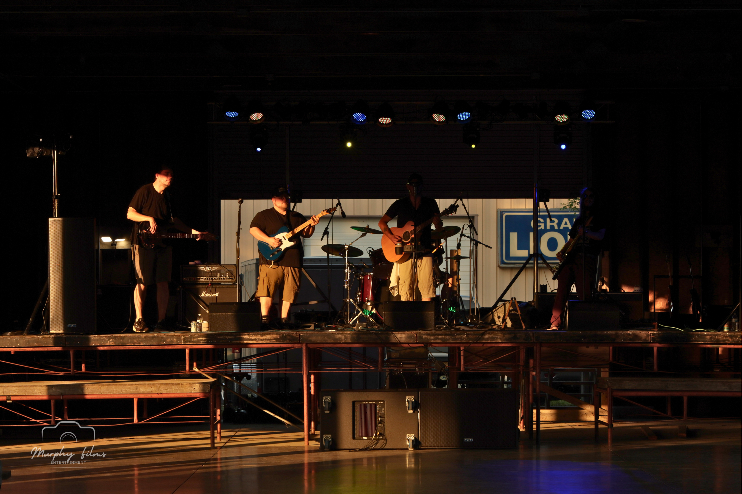 A band performs on an outdoor stage with four members playing guitars and drums, backlit by stage lights at sunset.