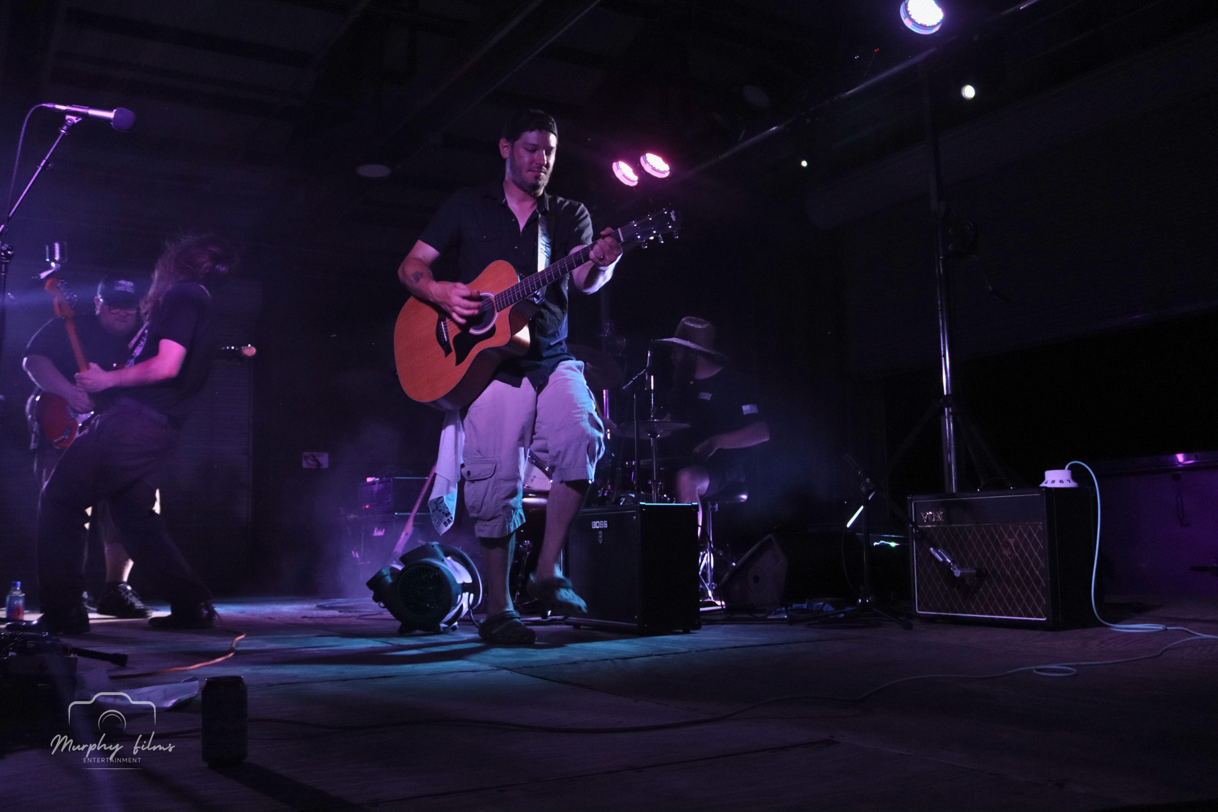 Musicians performing live on stage, with a guitarist prominently in the center, a keyboardist in the background, and a bassist on the left, under purple and pink stage lights.