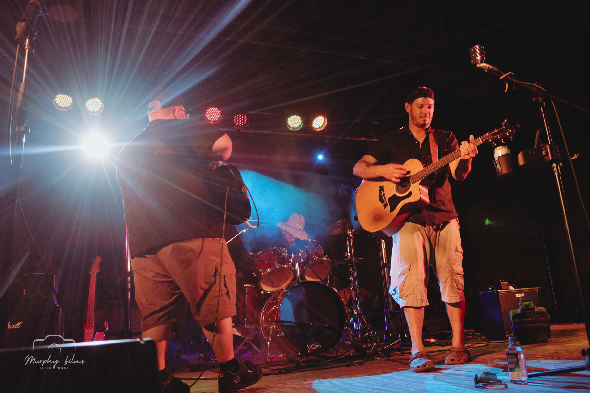 Three musicians perform on stage with stage lights, one playing an acoustic guitar, another with an electric guitar, and a drummer in the background.