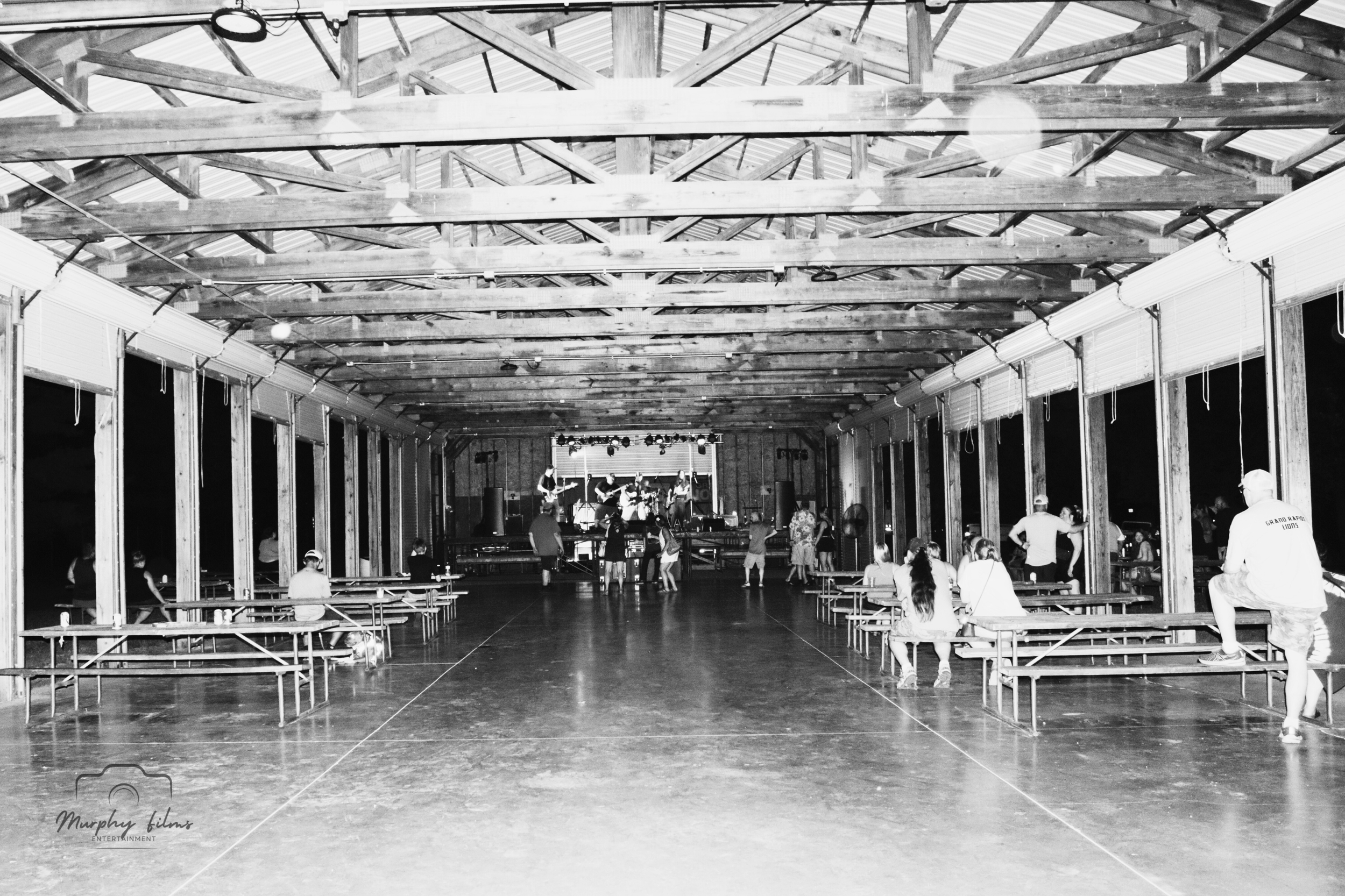 Black and white photo of an indoor event space with a stage at the far end, people sitting on benches along the sides, some standing or walking near the stage, wooden beams and rafters on the ceiling, and large windows along the sides.