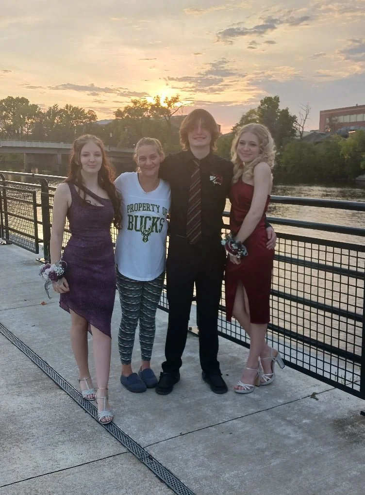 Four teenagers standing together outdoors during sunset, by a river, with trees and buildings in the background. Two girls are in dresses, one girl is in casual clothing, and one boy in a shirt and tie.