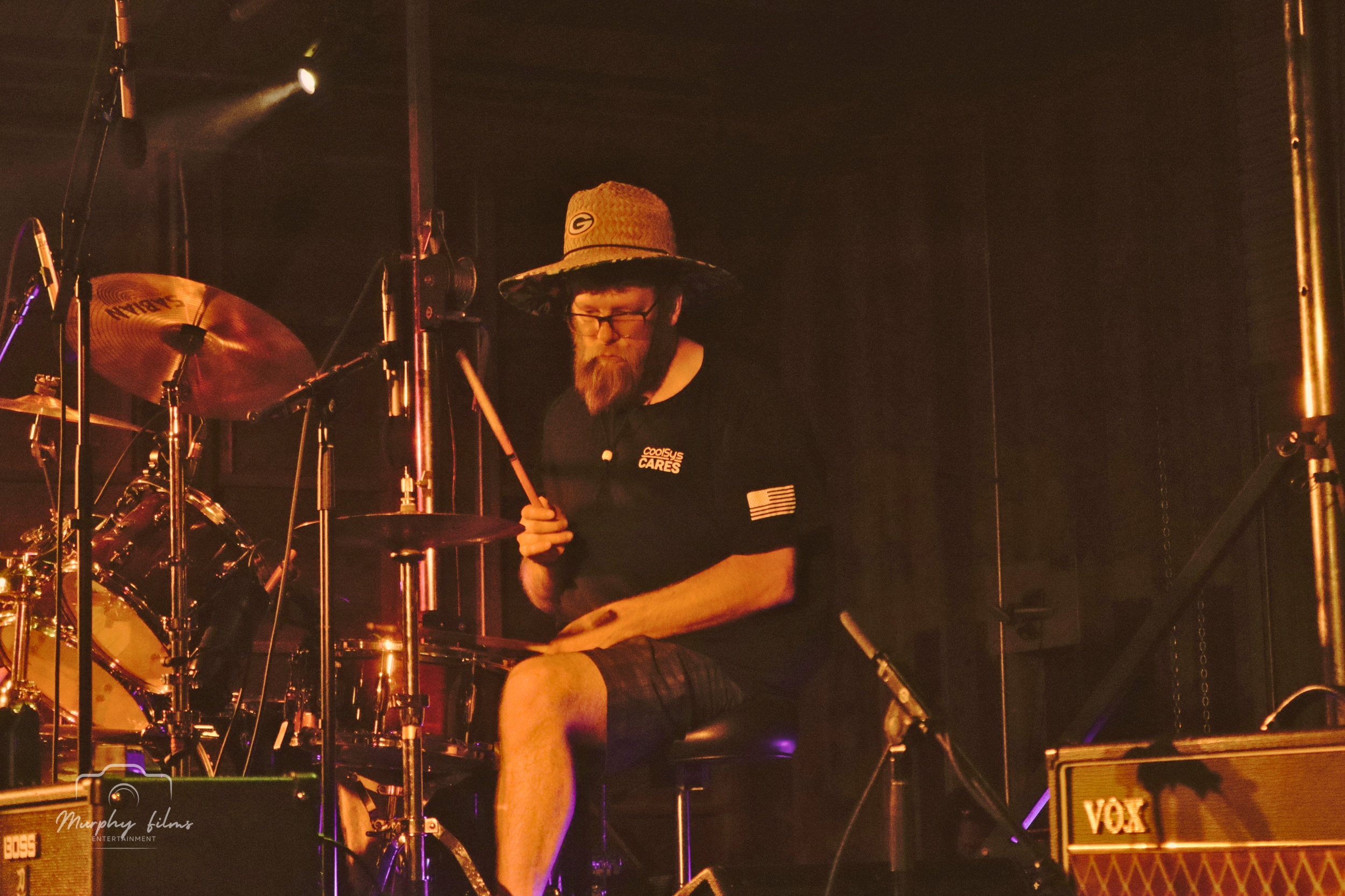 A man wearing a straw hat, glasses, and a black shirt with patches sits behind a drum set, performing on stage with dark background and stage lights.