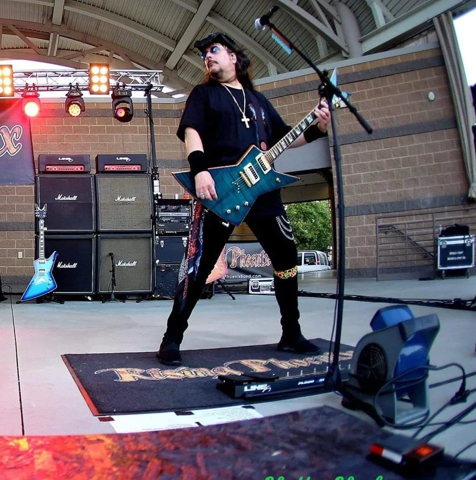 Male guitarist performing on stage with amplifier stacks, wearing sunglasses, a cross necklace, and dark clothing, under a partially open metal roof during daylight.