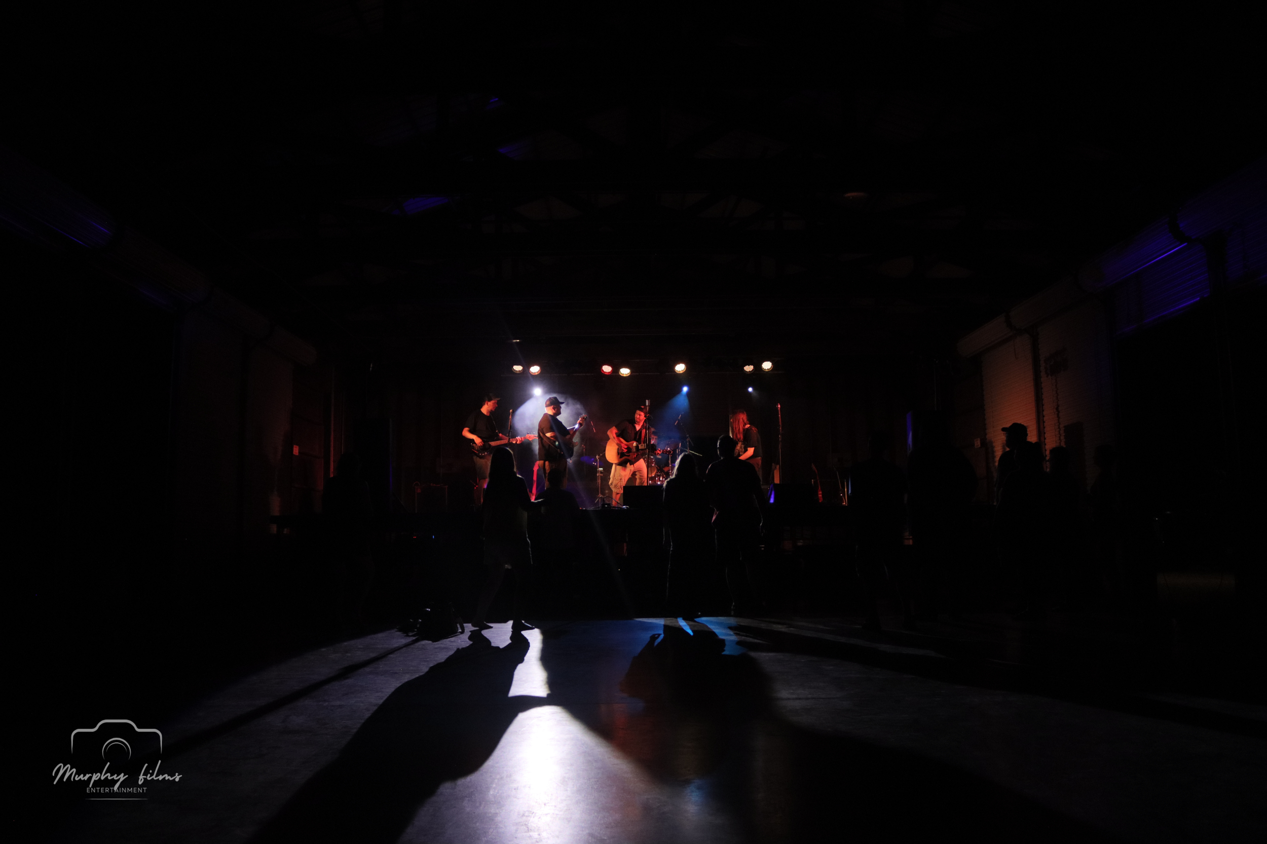 Silhouette of people dancing in front of a stage with musicians performing under stage lights at a concert or event.