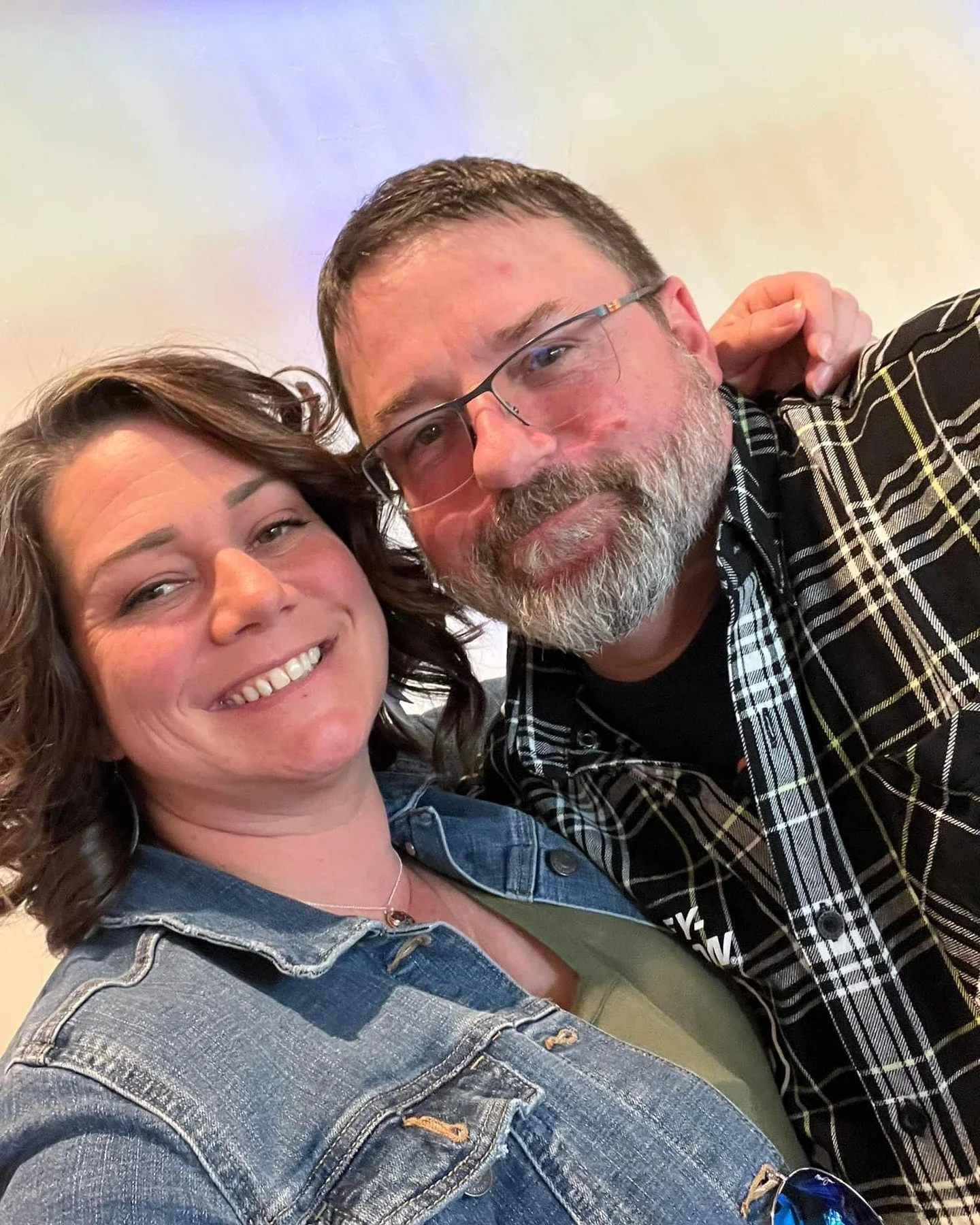 A woman and a man smiling for a selfie. The woman has wavy brown hair, and the man has short dark hair and a gray beard. They are close together, and the man is wearing glasses and a plaid shirt.