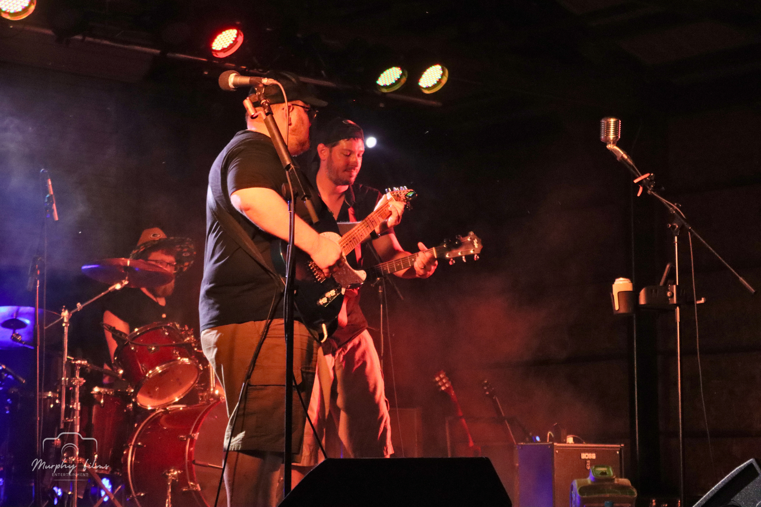 Two musicians playing guitars and a drummer on stage during a live performance, with stage lights overhead.