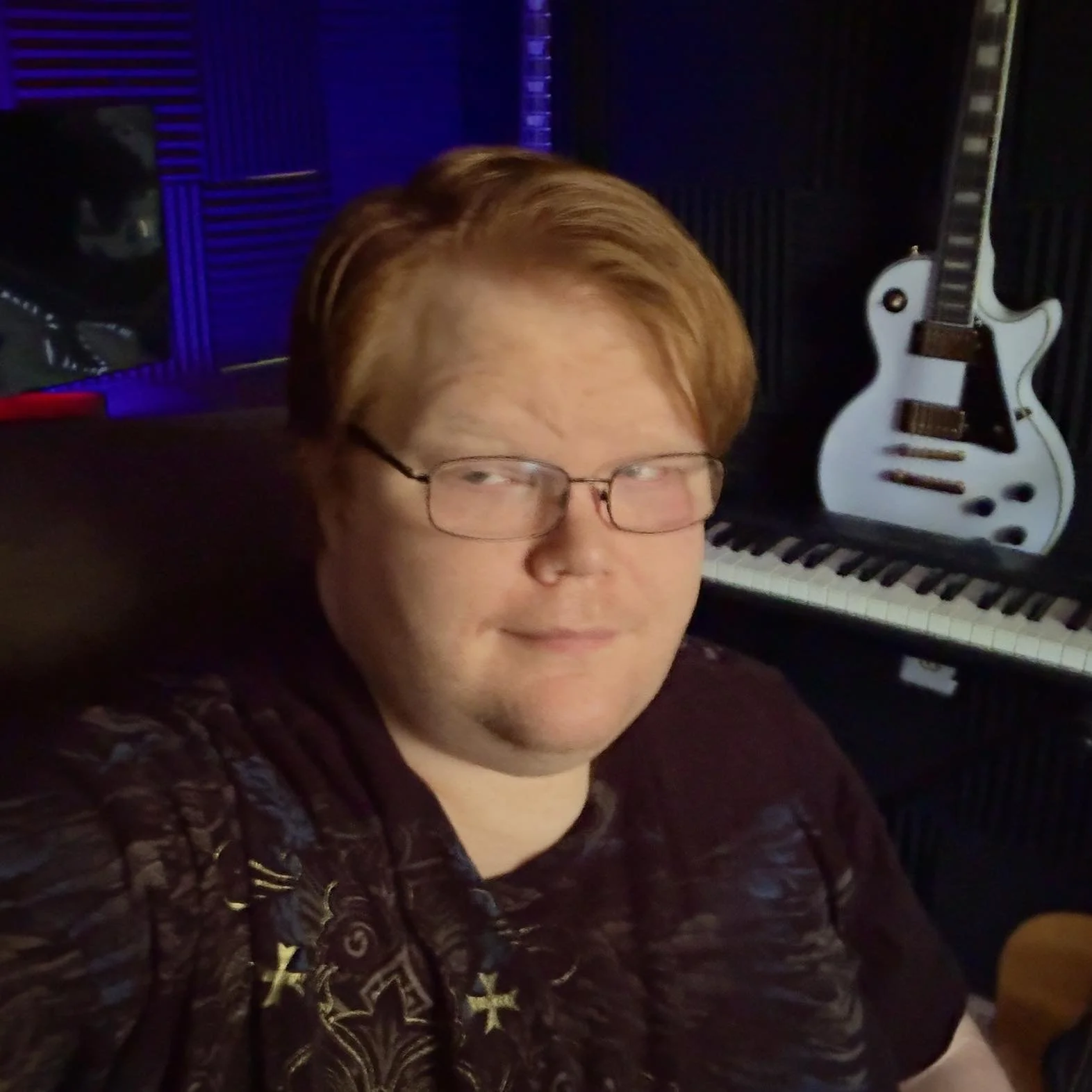 A man with reddish hair and glasses sitting in a music studio with a keyboard, a guitar, and soundproofing foam in the background.