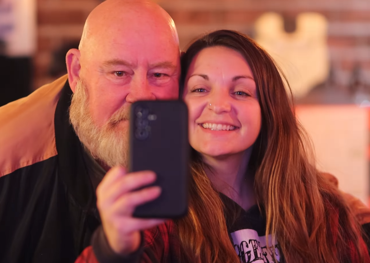 A man with a beard and a woman with long reddish hair taking a selfie together in a cozy, warmly lit setting.