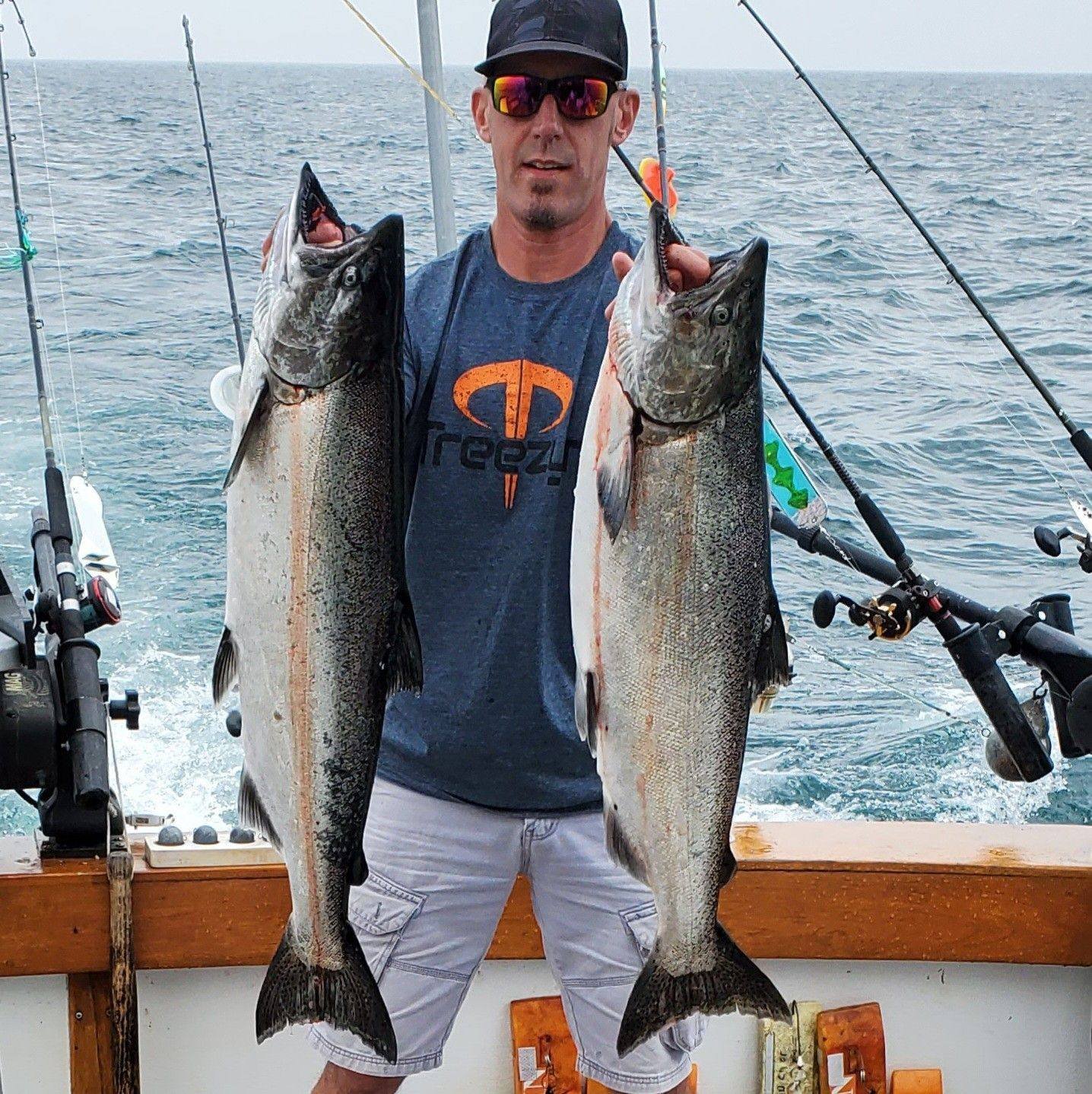 Man wearing sunglasses and a dark T-shirt holding two large fish aboard a boat on open water.